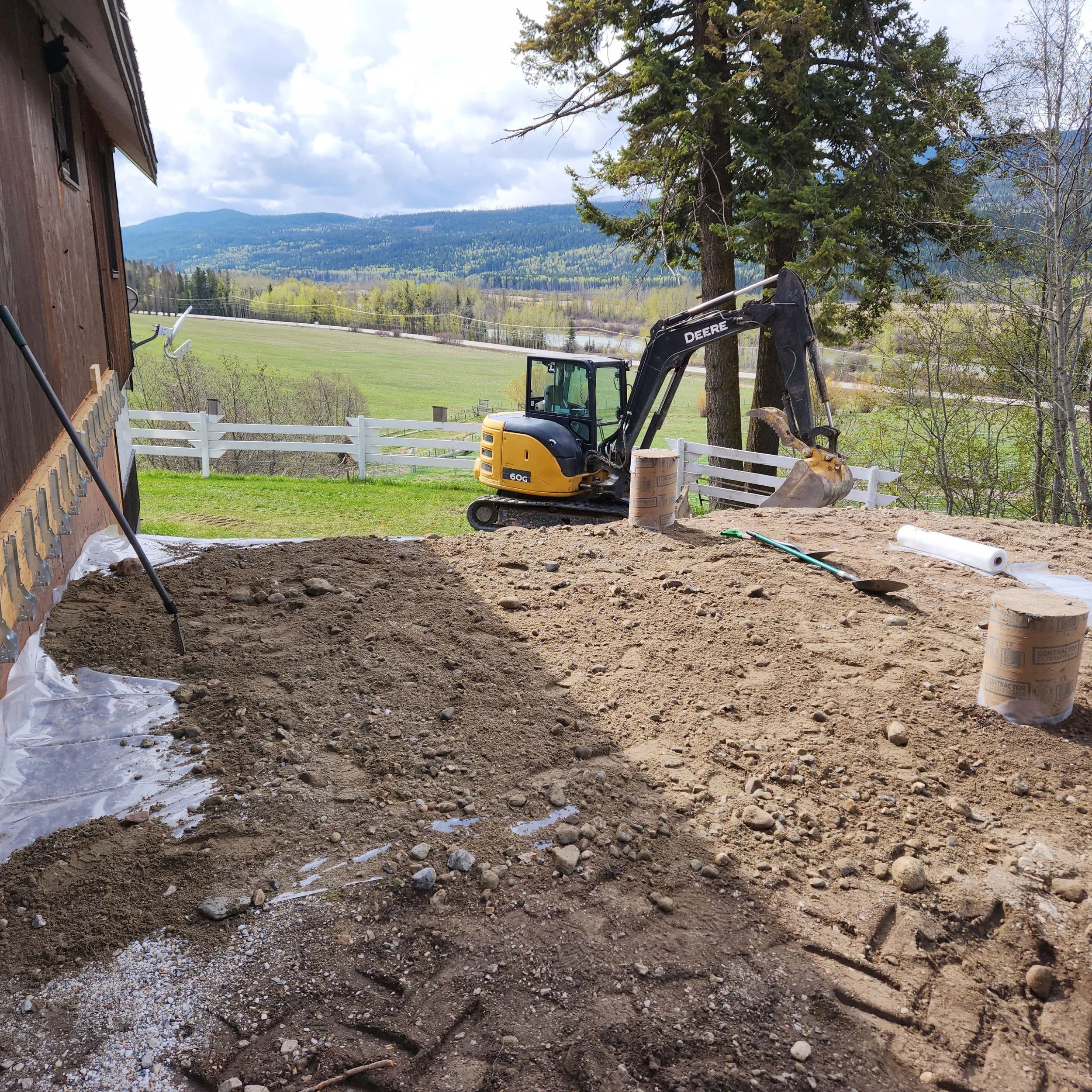 A construction site with excavated dirt, a Small John Deere mini excavator, and a white fence; in the background, there are trees, a grassy field, and mountains under a partly cloudy sky.