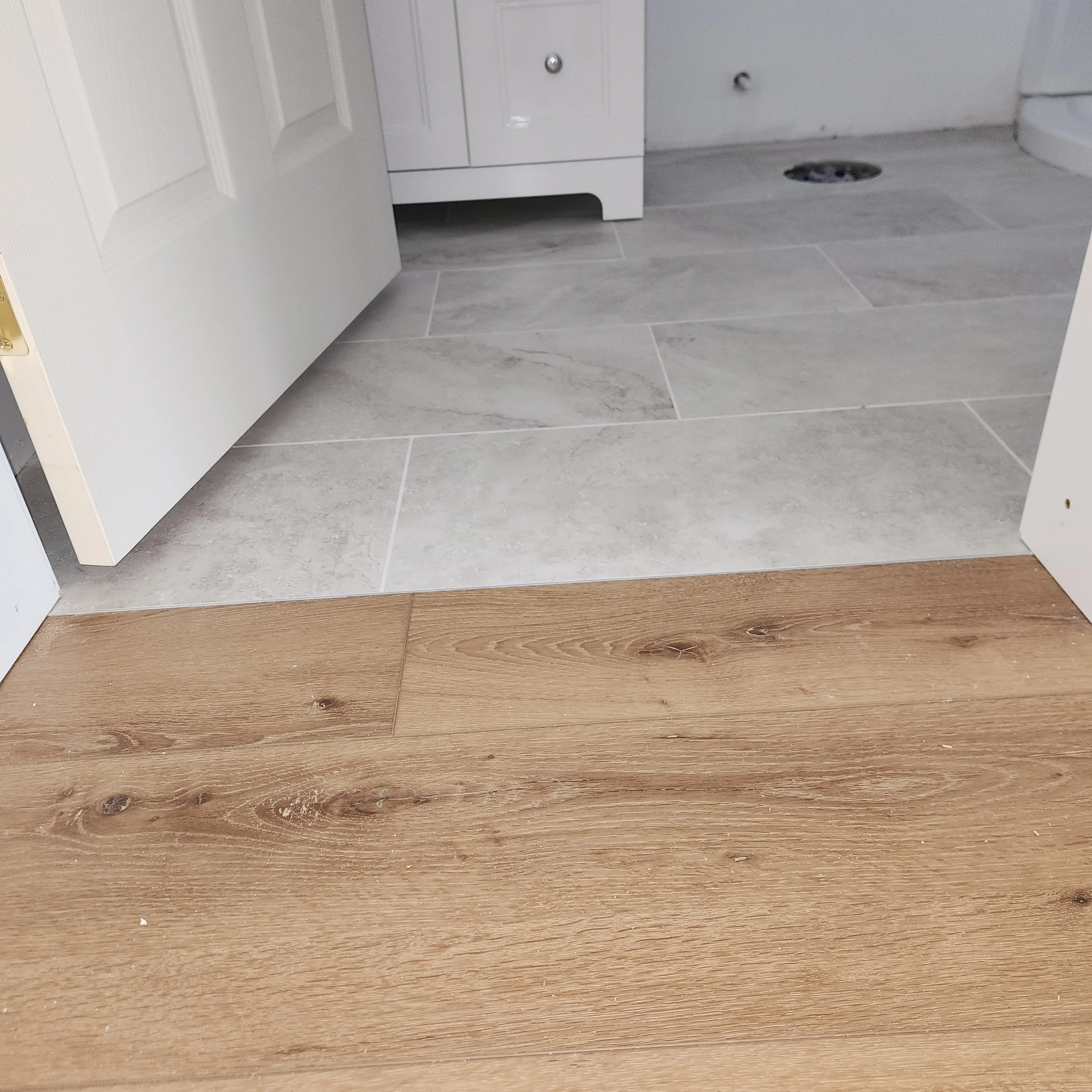View of a bathroom floor showing gray tiles and a small section of wooden flooring, with a white door partially open and a white cabinet or toilet base visible.