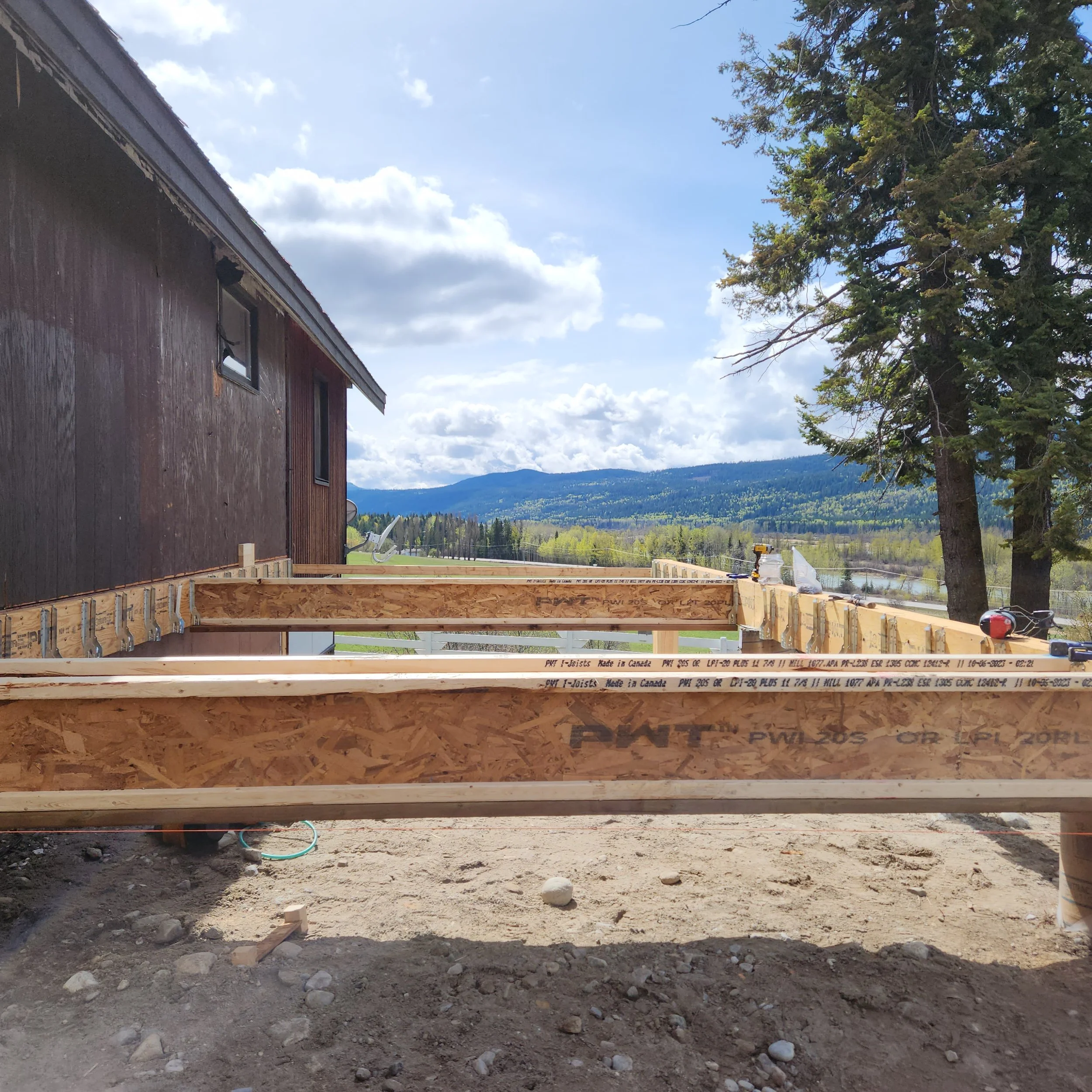 Construction site with wooden framing and dirt ground, next to a house with a mountain in the background and a large pine tree on the right.
