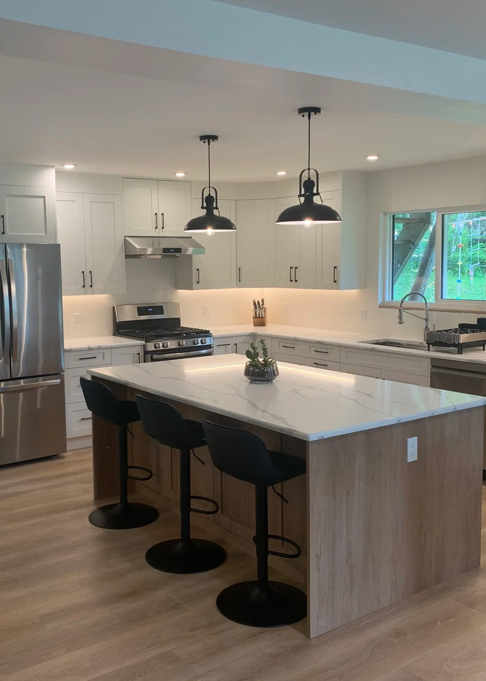 A modern kitchen with white cabinets, stainless steel appliances, and a large white marble island with wooden sides. Three black bar stools are positioned at the island. The kitchen has a window overlooking a backyard with greenery and a climbing fra