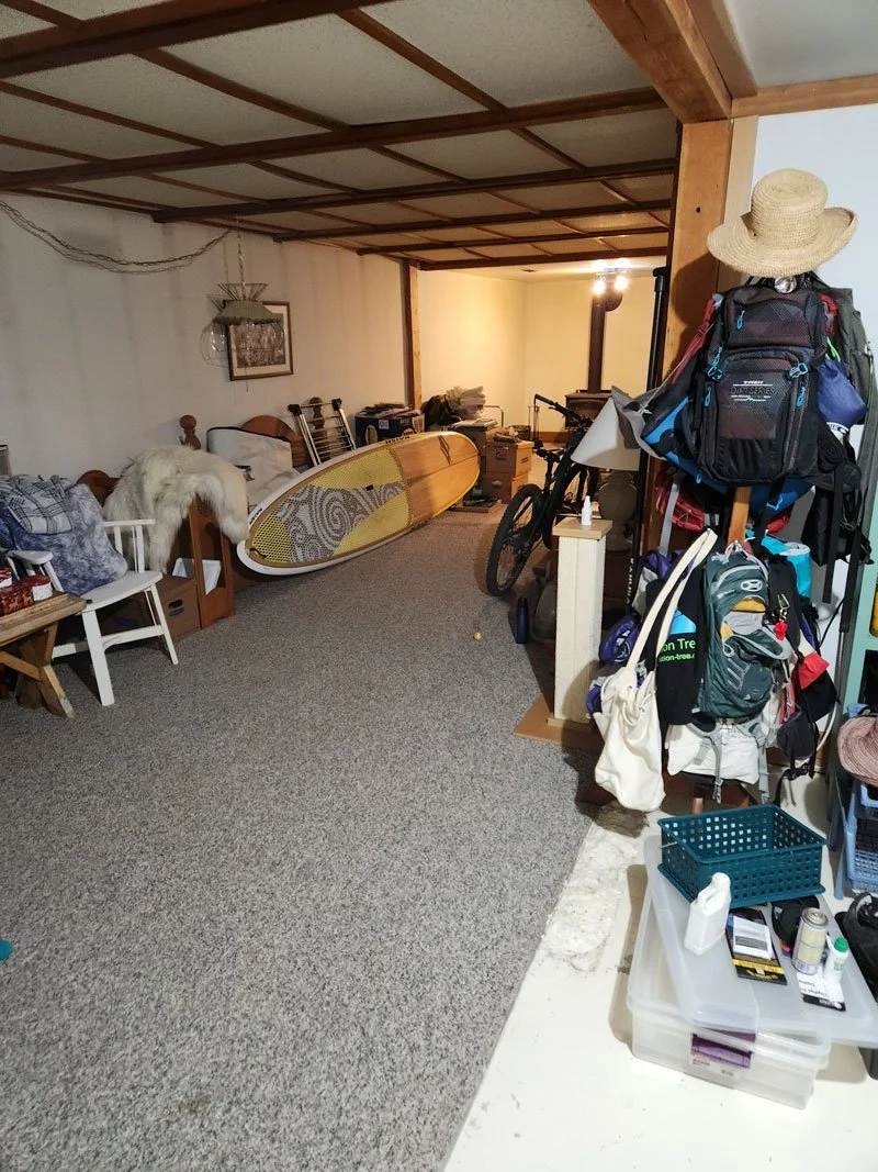 Living room or basement with a surfboard, bicycles, backpacks, and various household items and furniture.