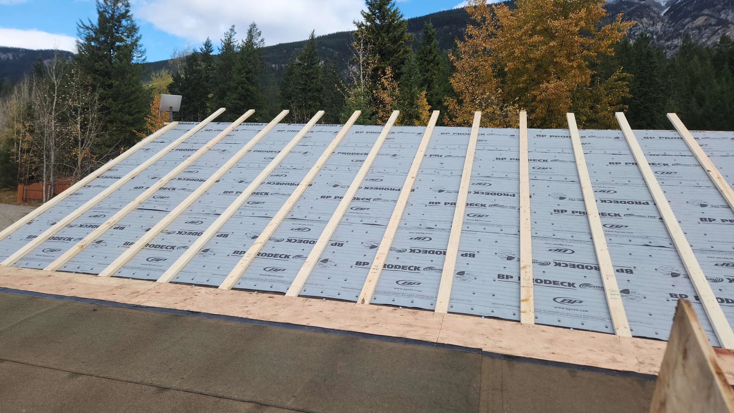 Roof under construction with wooden beams and underlayment, surrounded by trees and mountains in the background.