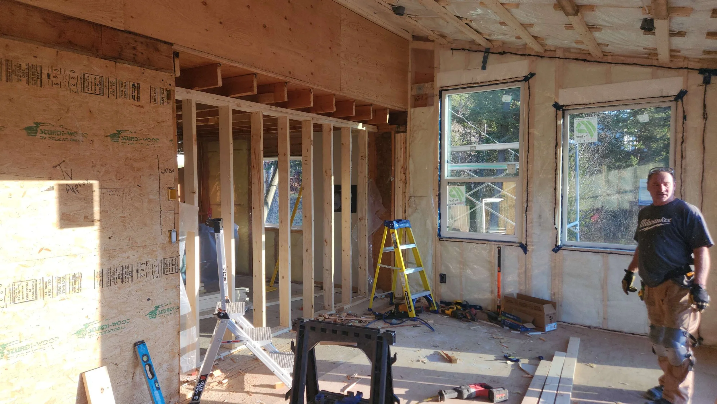 Interior of a house under construction with unfinished walls and exposed stud framing, two windows installed, a yellow step ladder, and scattered tools. A man wearing a Milwaukee t-shirt is walking through the space.