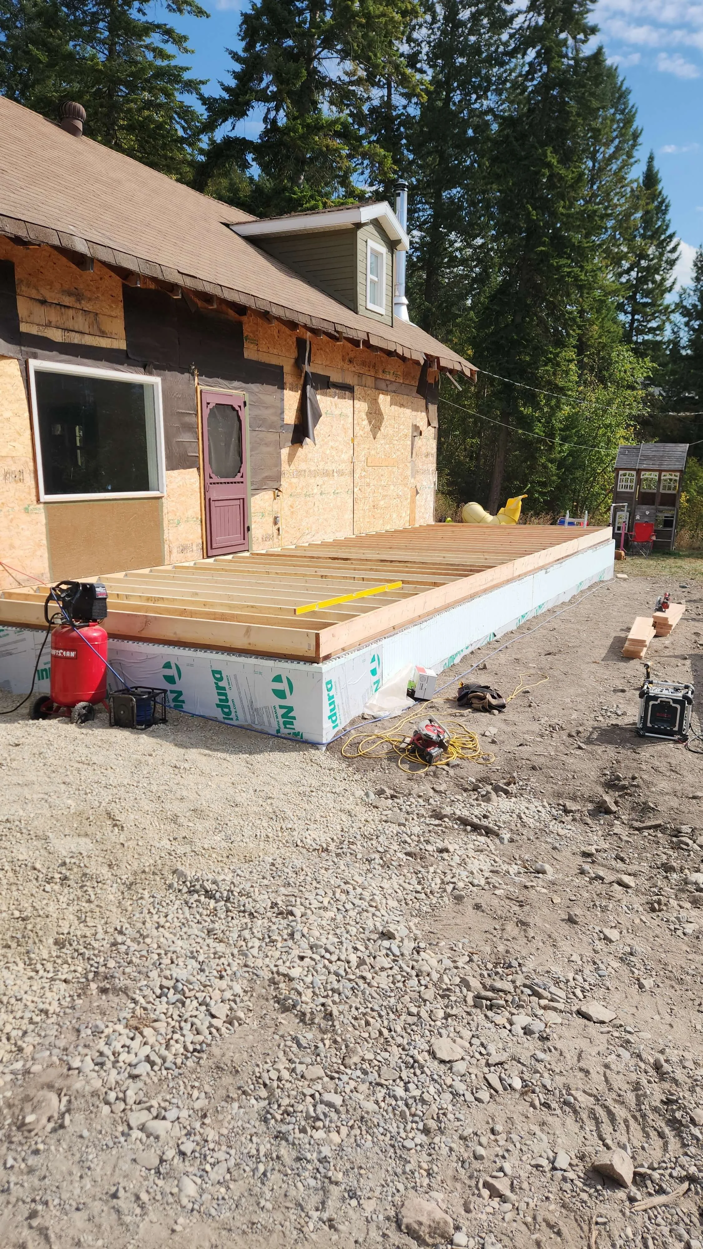 Construction site with a house in progress showing an unfinished deck frame, power tools, and construction materials on gravel ground, surrounded by trees.