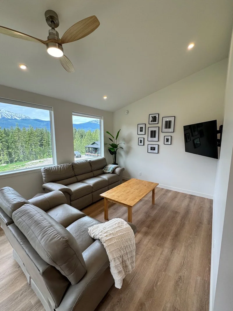 Living room with two leather sofas, wooden coffee table, large windows showing mountain and forest view, modern decor, mounted TV, ceiling fan, and a gallery wall of framed photos.