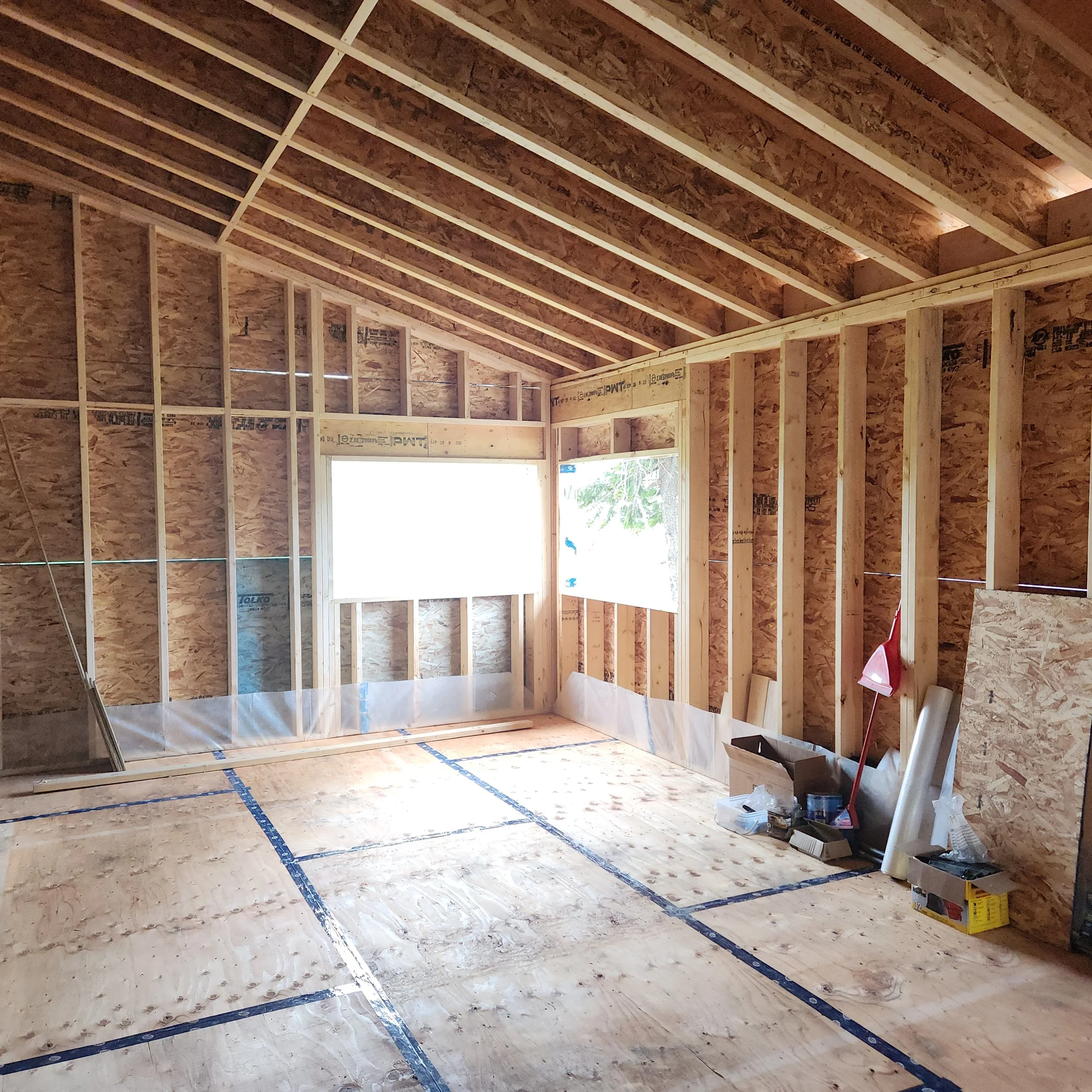 Interior of a house under construction with exposed wooden framing, unfinished walls, and a partially installed ceiling. Construction materials and tools are scattered on the floor.