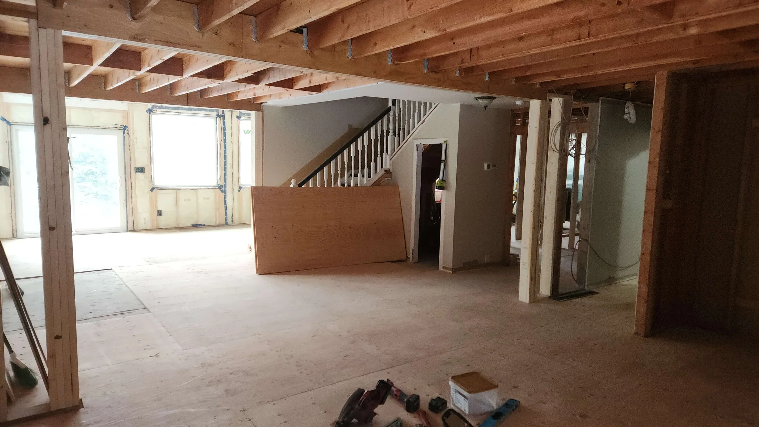 Interior view of a house under construction with exposed wooden beams, drywall framing, and unfinished walls. There is a staircase with a black handrail, multiple windows, construction tools, and materials visible.