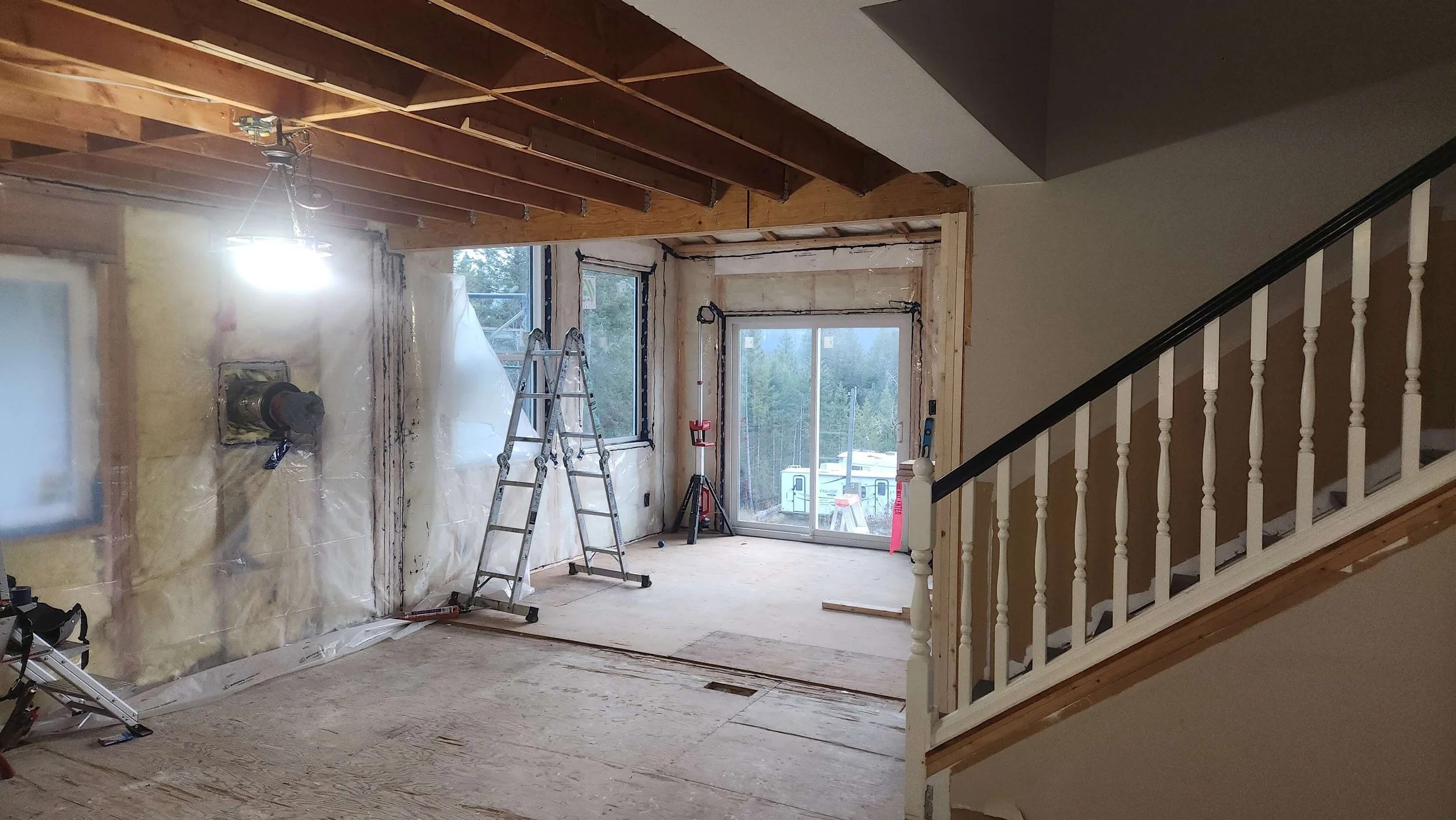 Interior of a house under renovation with exposed wooden ceiling beams, ladders, construction tools, and large windows providing natural light.