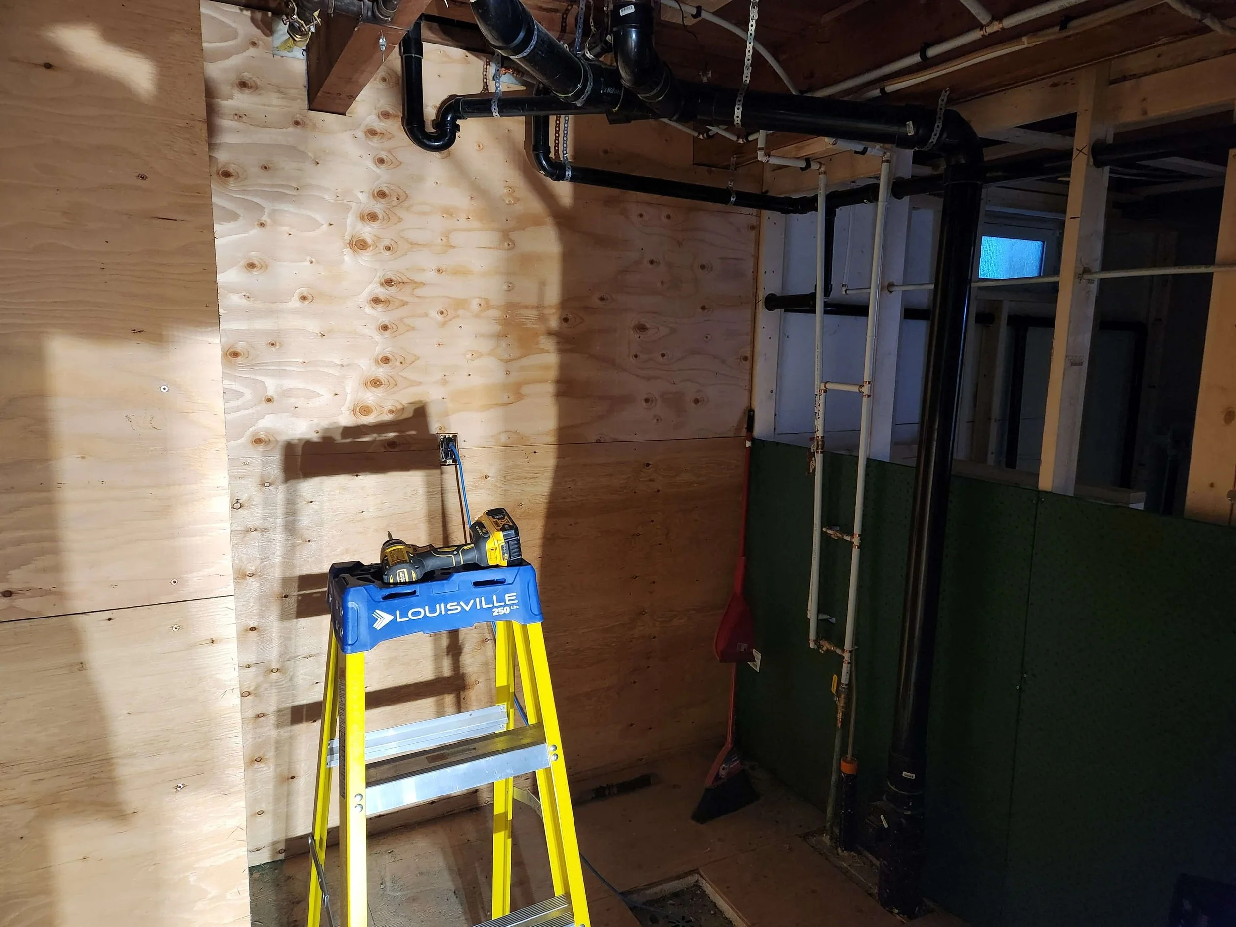 Construction site with partially built wooden wall, black plumbing pipes, and a yellow ladder with a toolbox on the floor.
