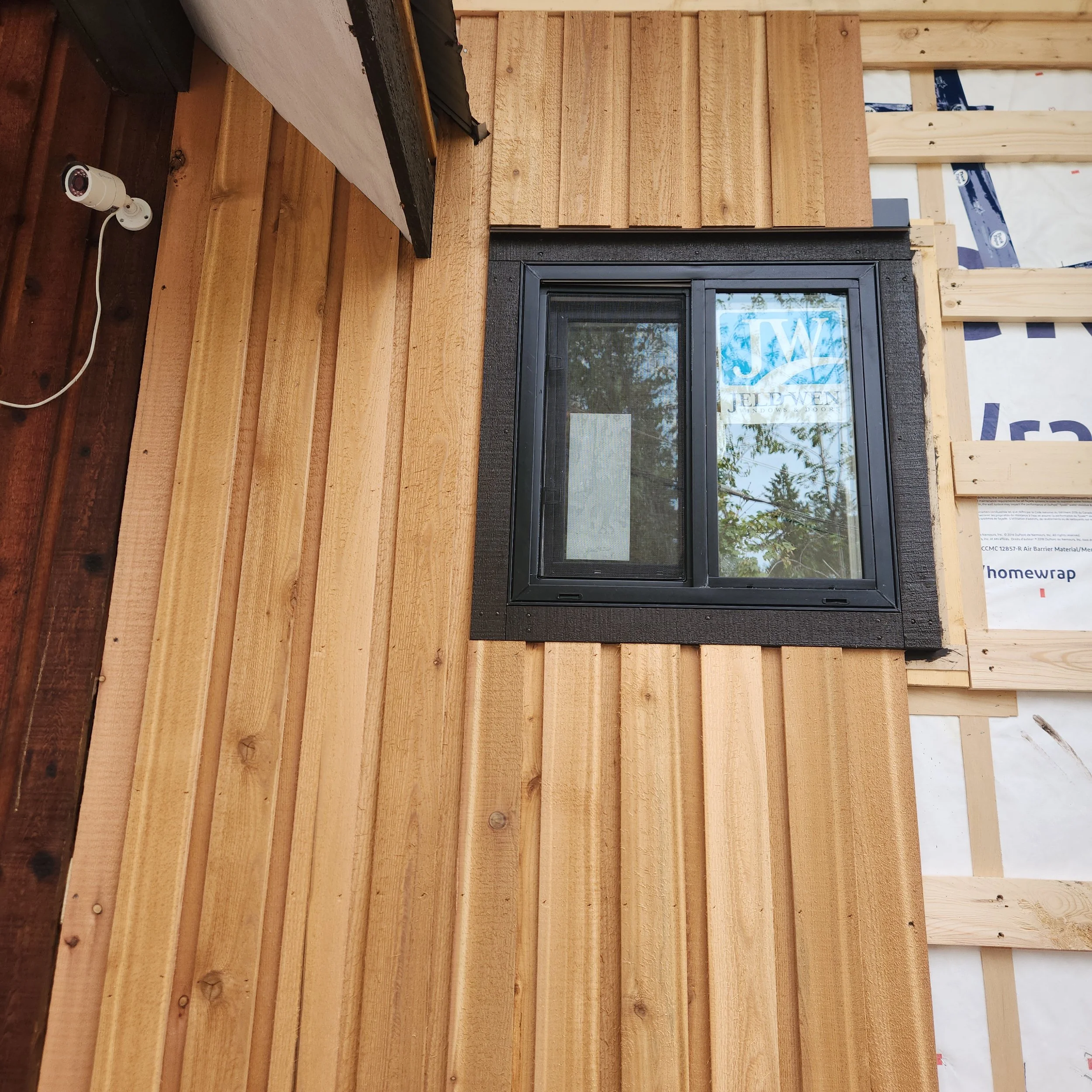 Photo of a wooden exterior wall under construction with a black-framed window and a security camera mounted on the wall.