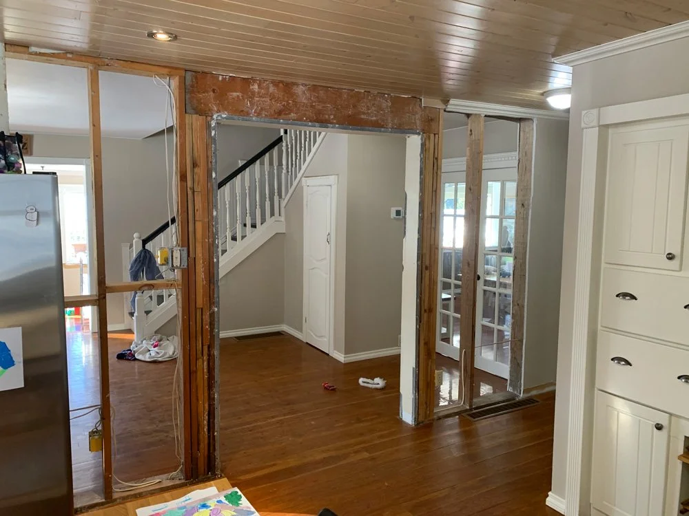 Kitchen with a partially removed wall, exposing wooden studs and electrical wiring, leading to a dining area with sliding glass doors.