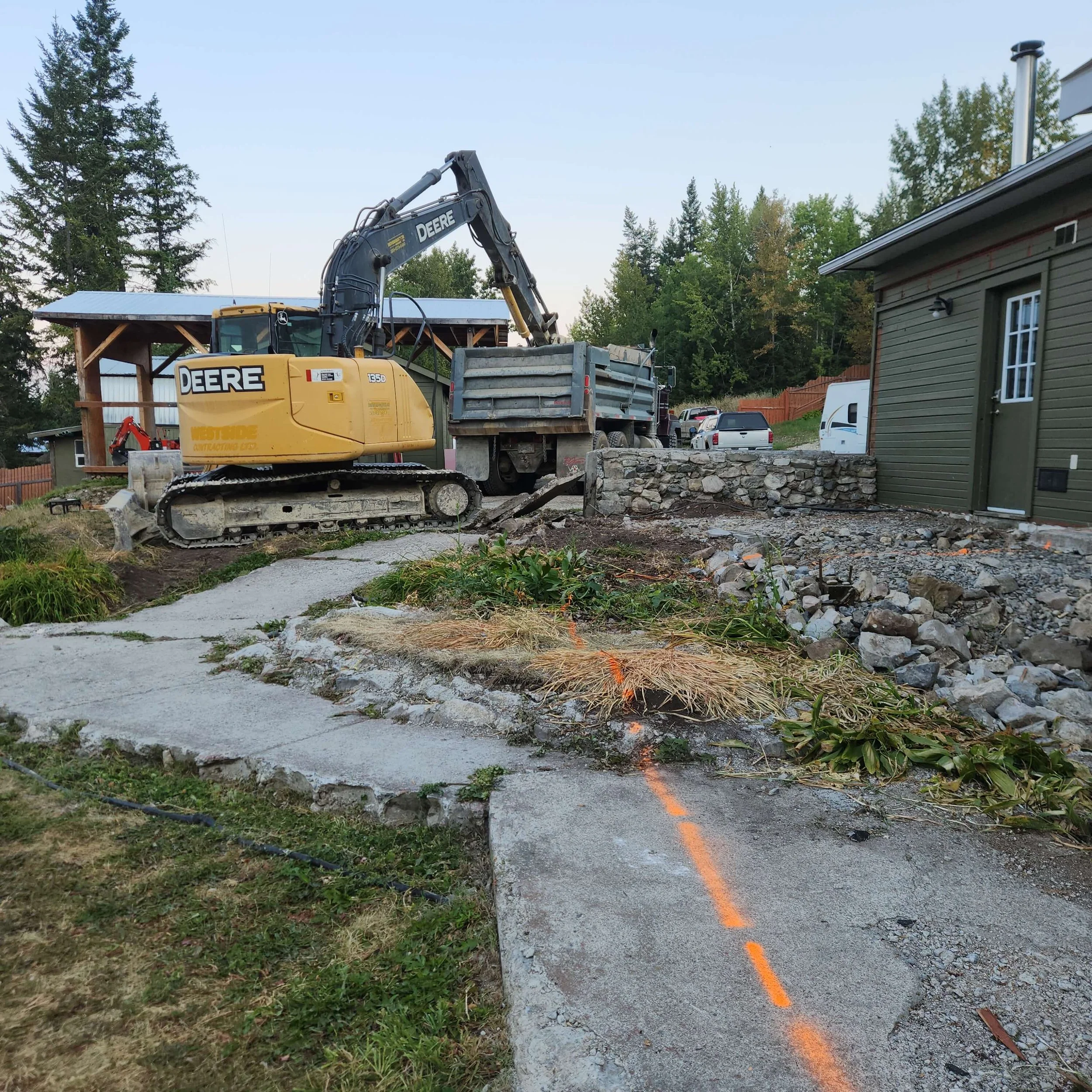 Construction site with a yellow John Deere excavator, a partially broken concrete sidewalk, a green house, and trees in the background.