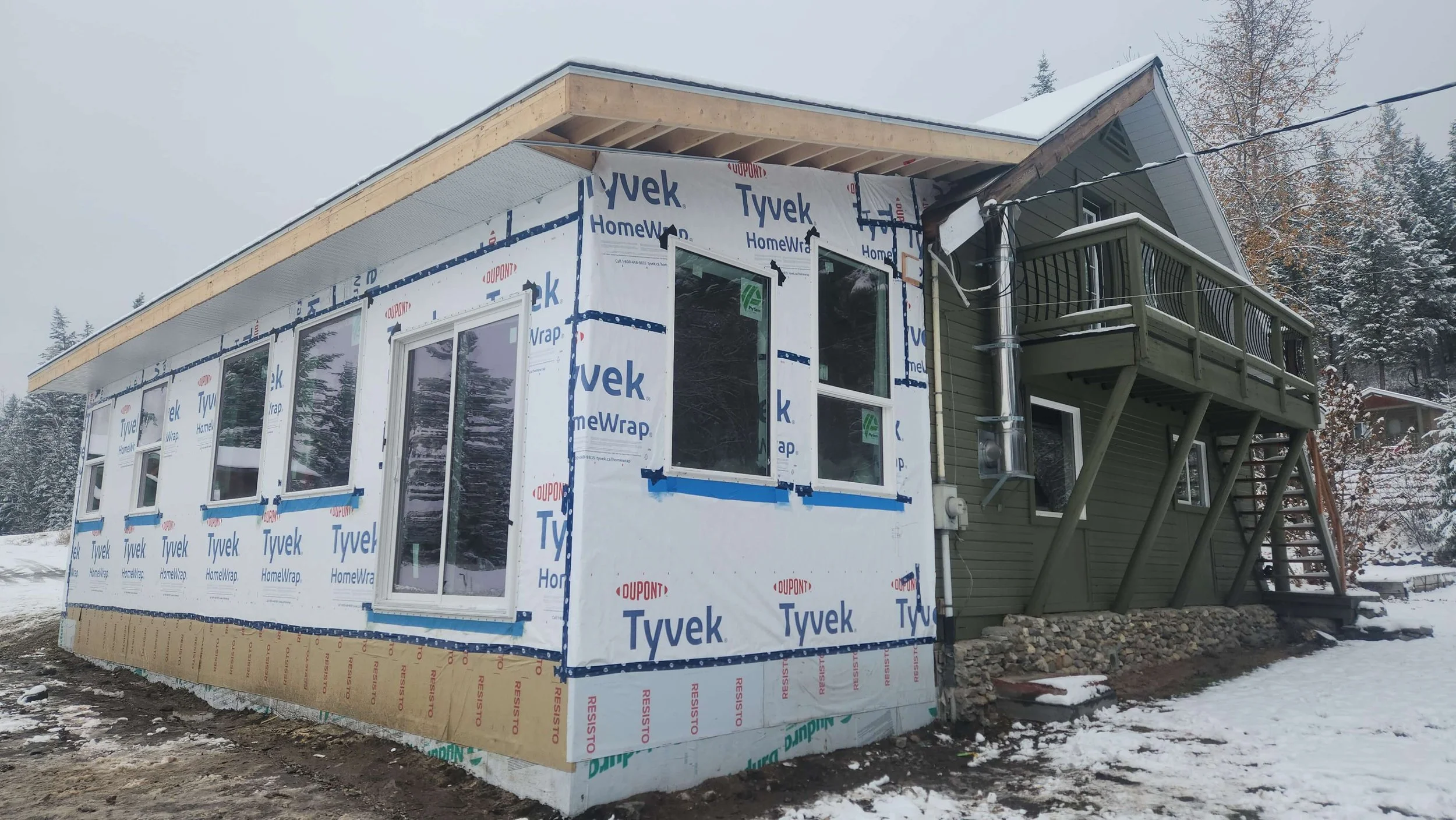 Side view of a house under construction with insulation and windows installed, and a small balcony on the upper floor, set in a snowy landscape.