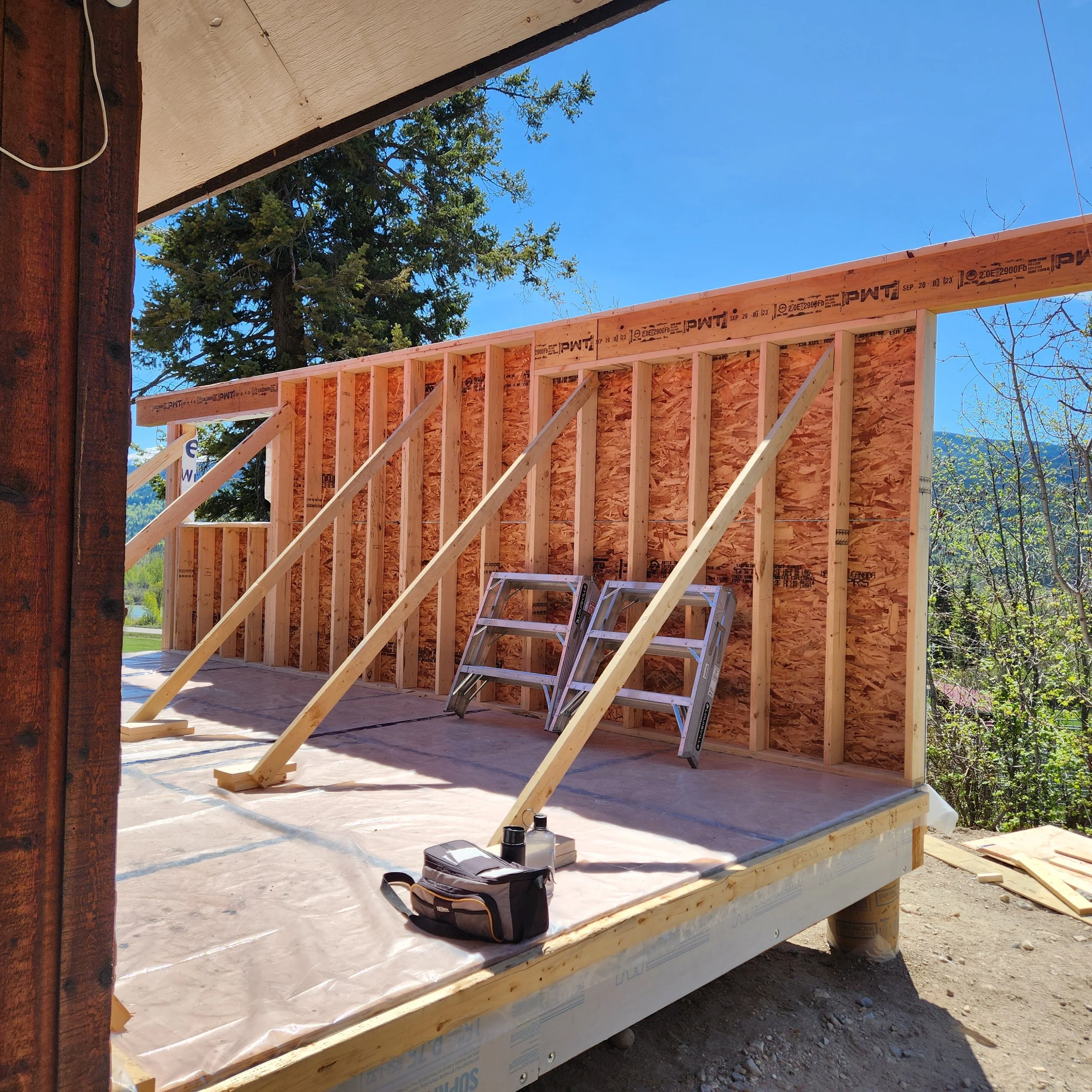 Construction site with wooden framing for a building, ladder leaning against the frame, and tools on the floor. Blue sky and trees in the background.
