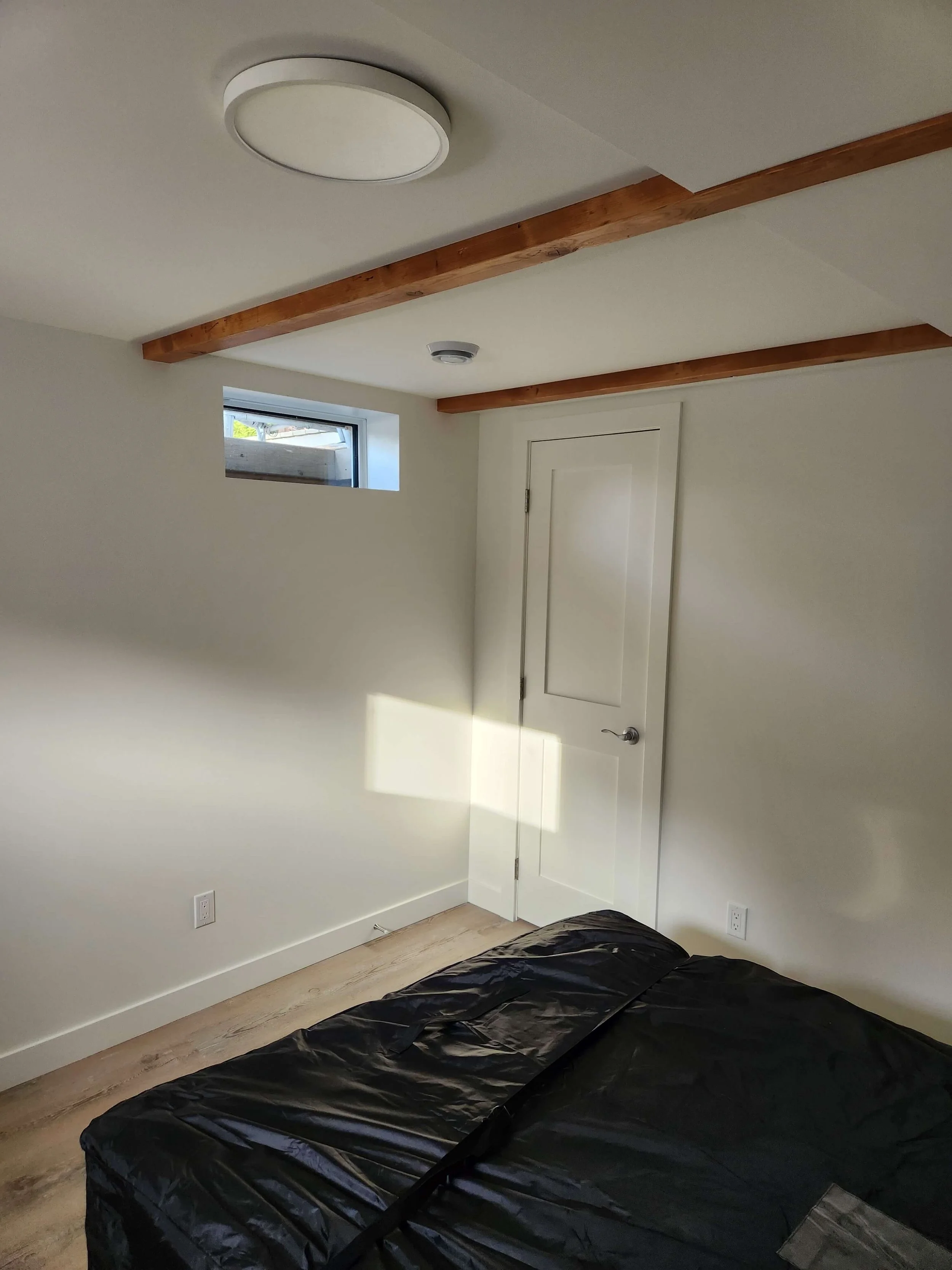 A room with a small rectangular window, white walls, a white door, wood flooring, a ceiling light, exposed wooden beams, and a black plastic-covered object on the floor.