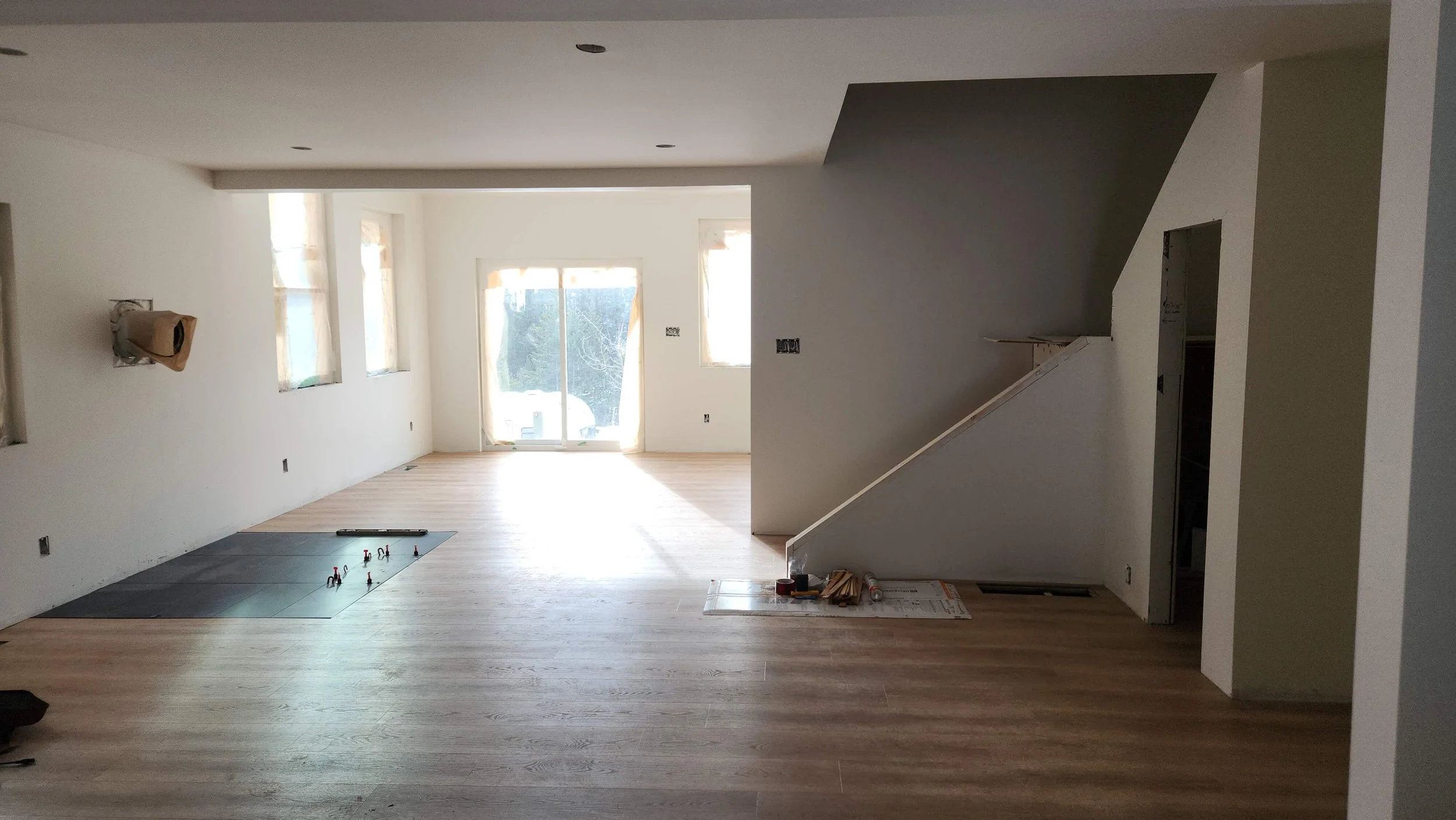 Interior of a house under construction with white walls, large windows, and a sliding door. The floor is partially finished with hardwood and some tiles. Construction materials and tools are scattered on the floor.
