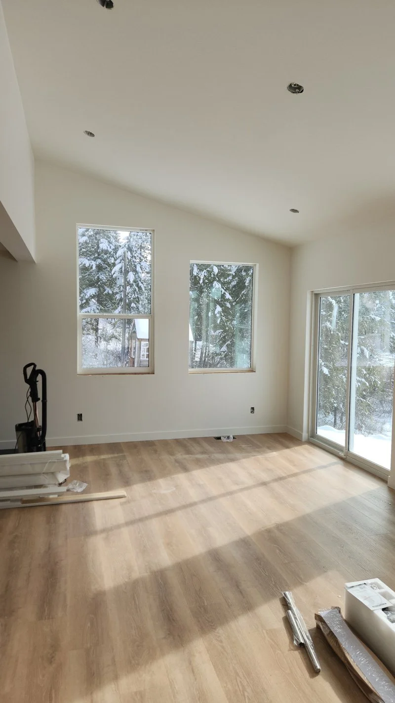 Empty living room with three large windows showing snow-covered trees outside, a sliding glass door, light-colored wooden flooring, and a white ceiling with open recessed light fixtures, under construction or renovation.