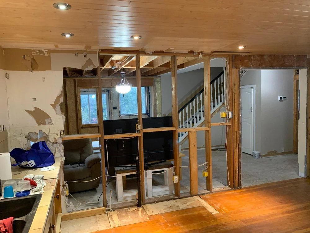 Home renovation in progress with exposed wall studs, unfinished drywall, and a view into the living room with staircase and a window.