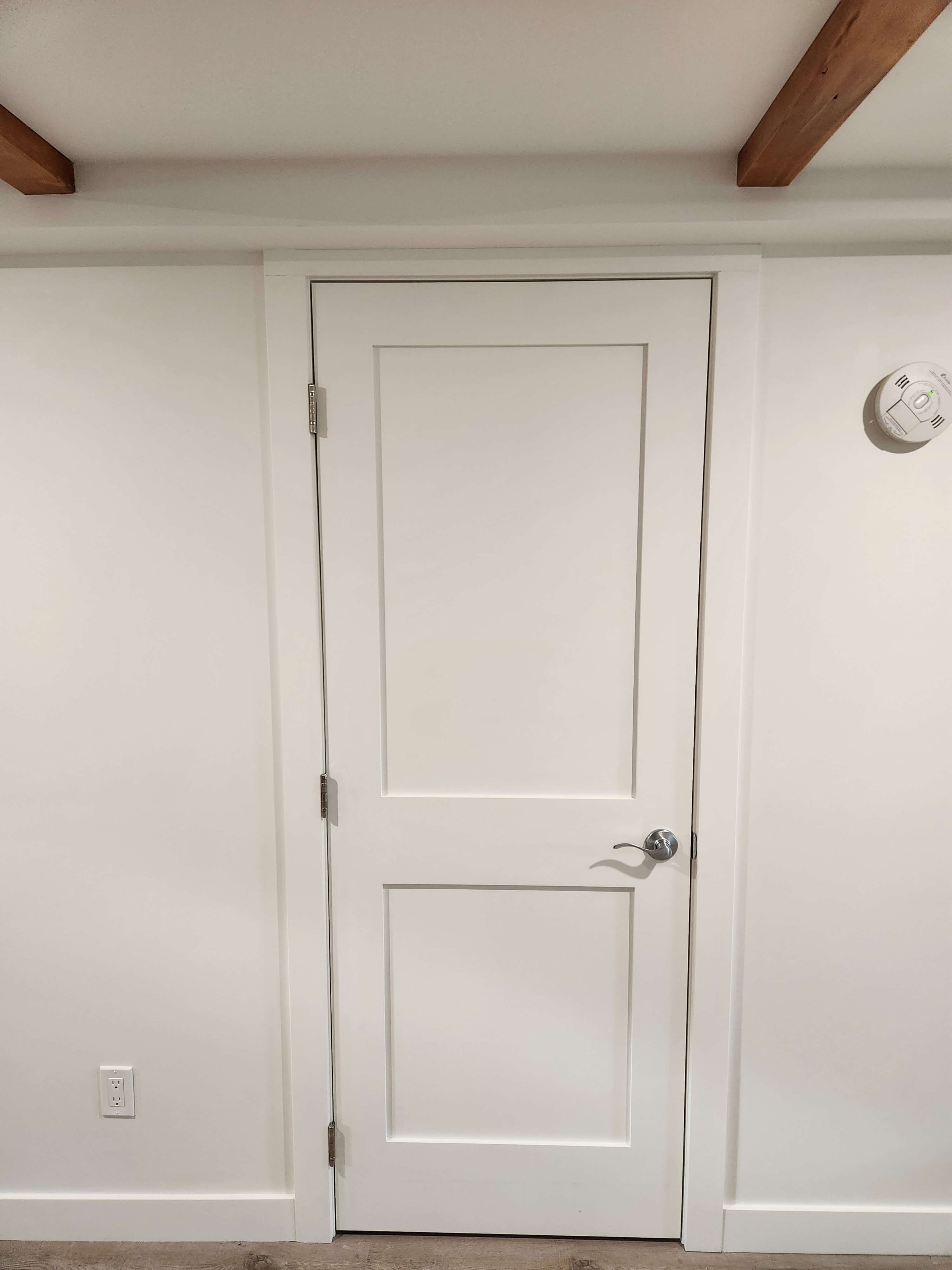 White interior door with a silver handle, set in a white wall with a wooden ceiling beam visible at the top and a smoke detector on the right wall, with a power outlet near the floor.