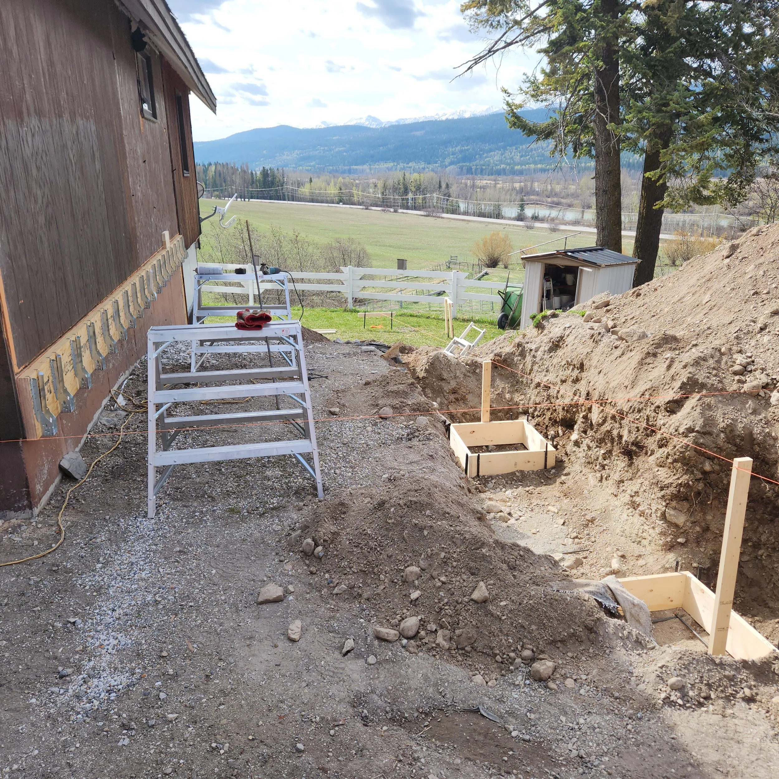 A construction site with dirt and gravel, a partially built house wall with exposed sheathing, tools, and equipment scattered around, trees, a mountain range with snow-capped peaks in the background, and a partly cloudy sky.