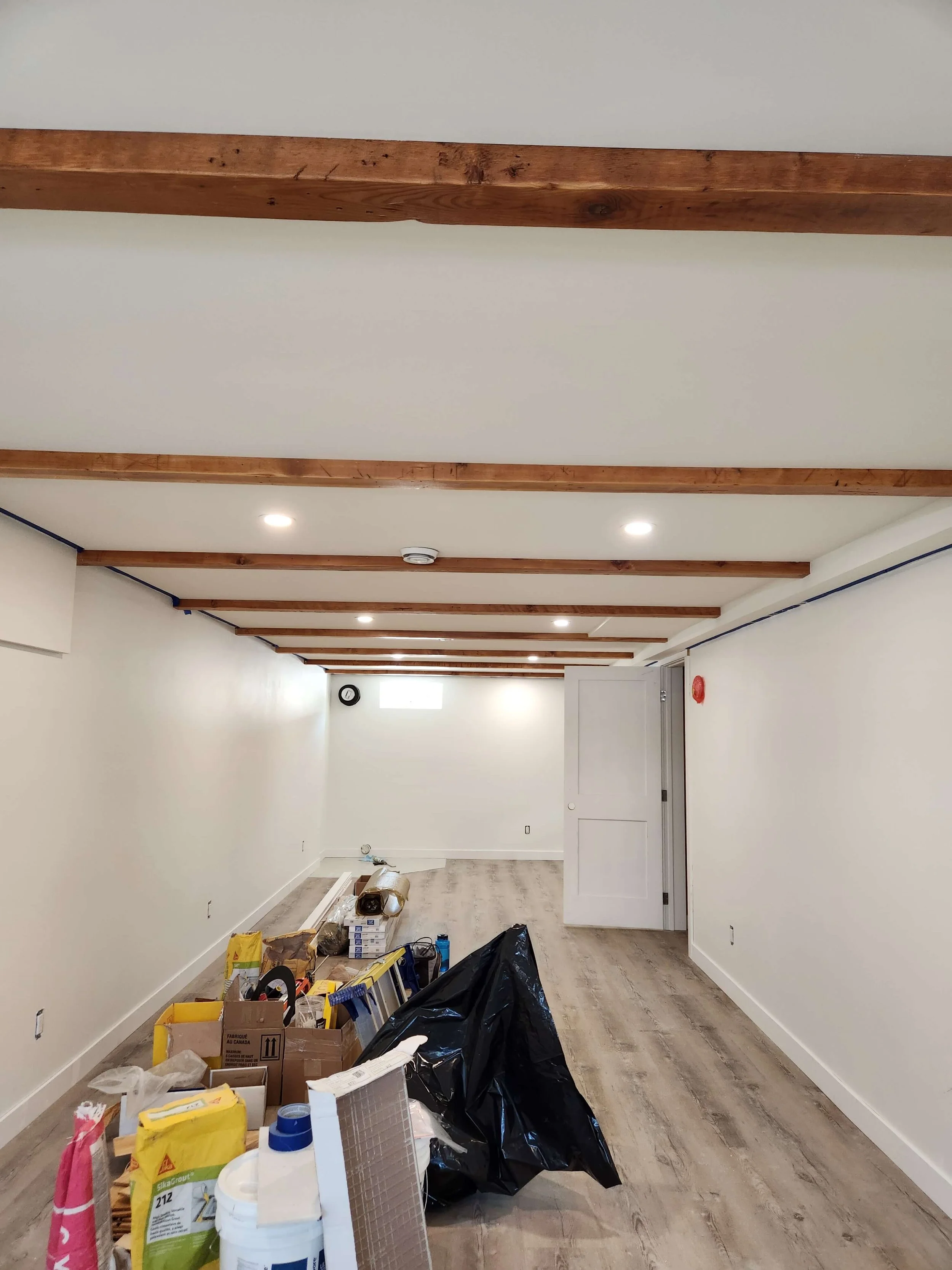 Empty hallway with wooden beams on the ceiling, construction materials, packages, and bags on the floor, room appears to be under renovation.