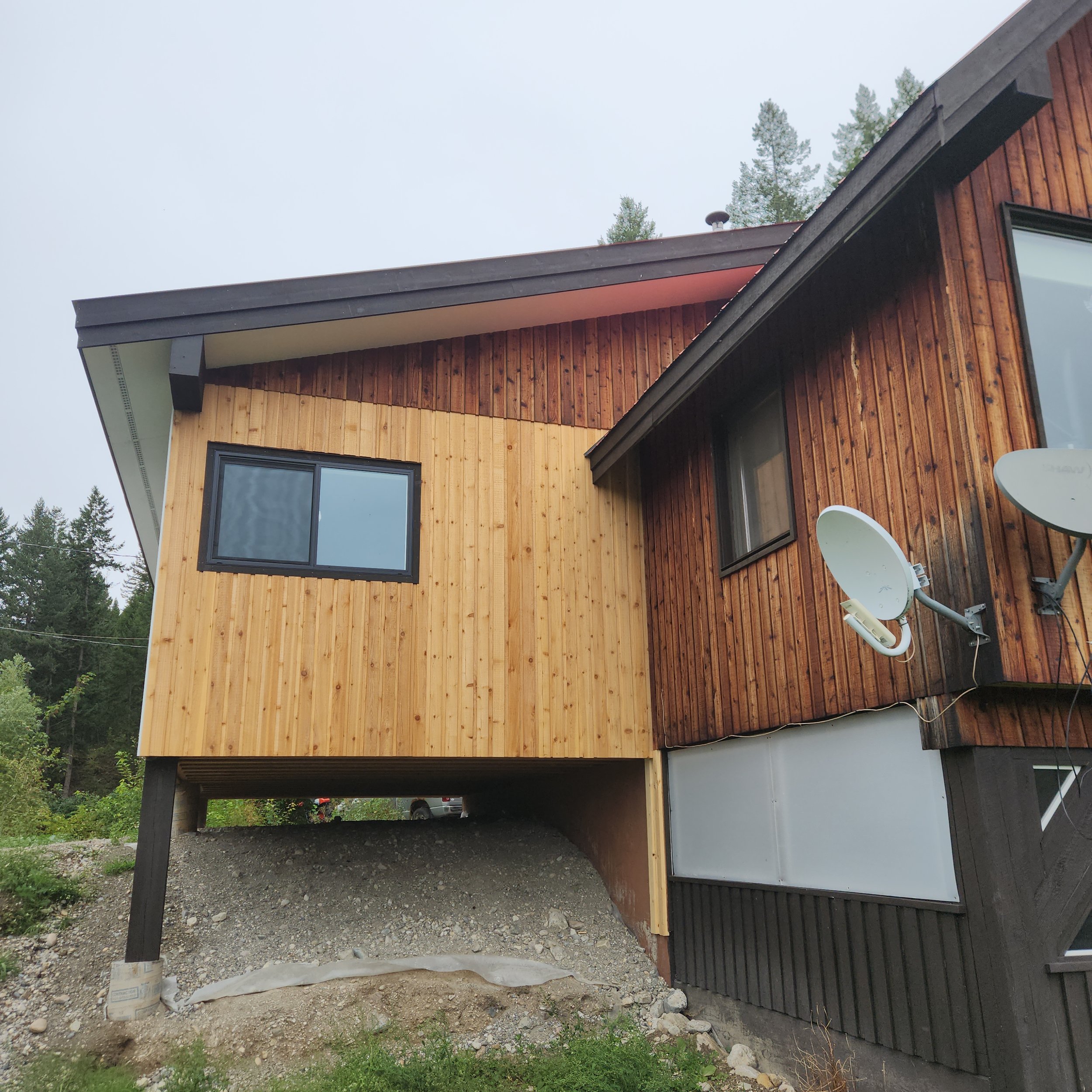 Construction site showing a modern house with wooden exterior, large windows, and an overhang supported by black columns; some construction materials and dirt are visible at the site.