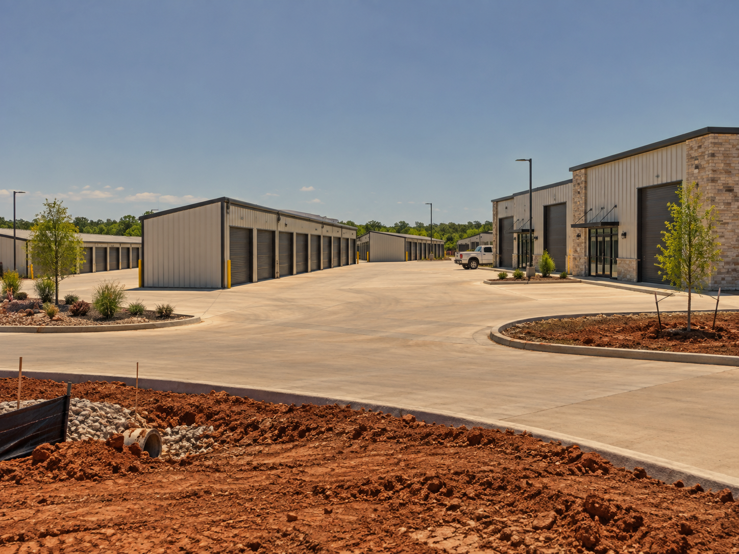 A newly constructed commercial storage facility with multiple metal unit buildings, a concrete driveway, and landscaped areas with small trees and shrubs, under a clear blue sky.