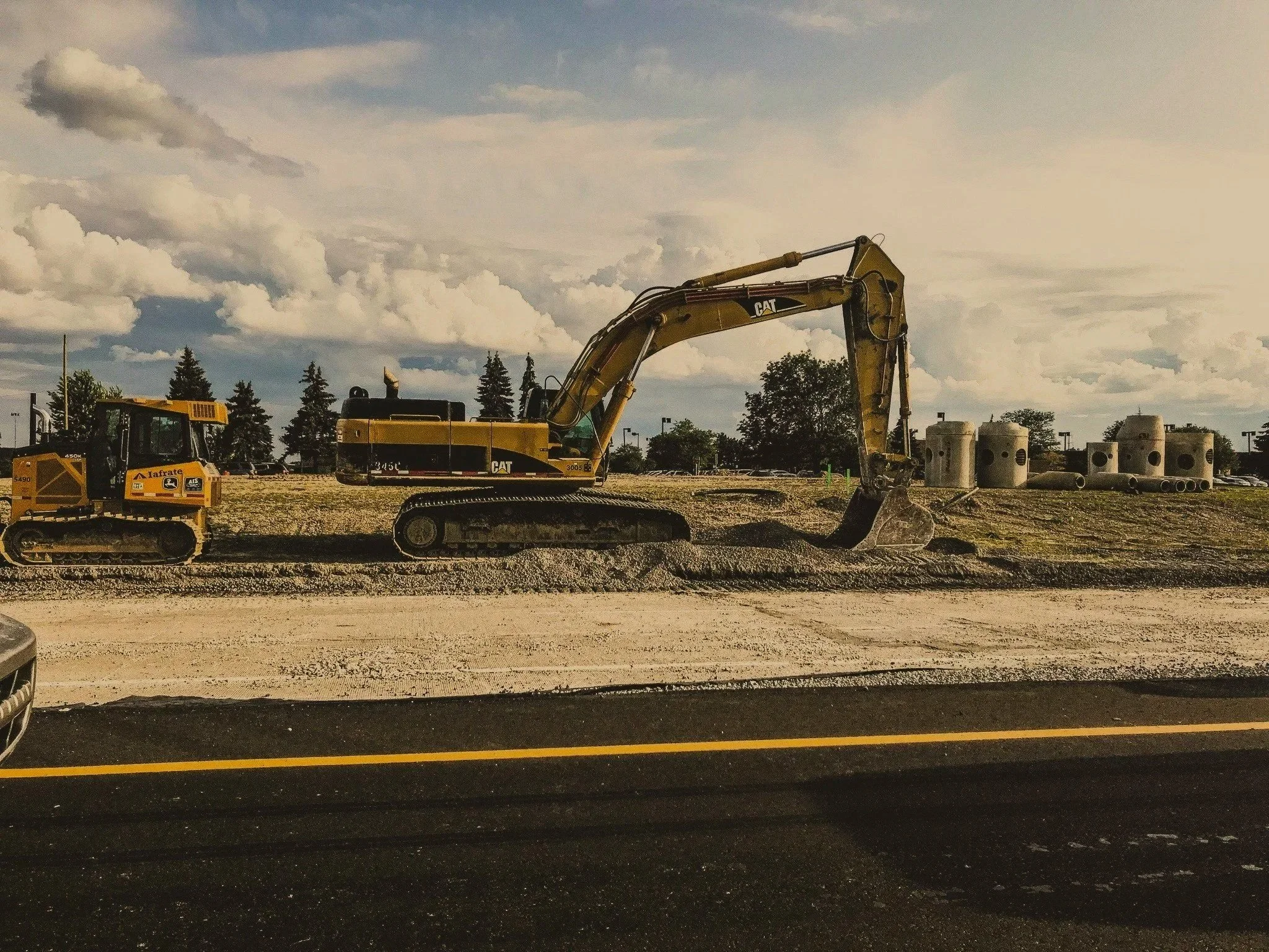 A construction site with a yellow Caterpillar excavator and a yellow John Deere bulldozer parked on a dirt ground, with trees and a cloudy sky in the background.