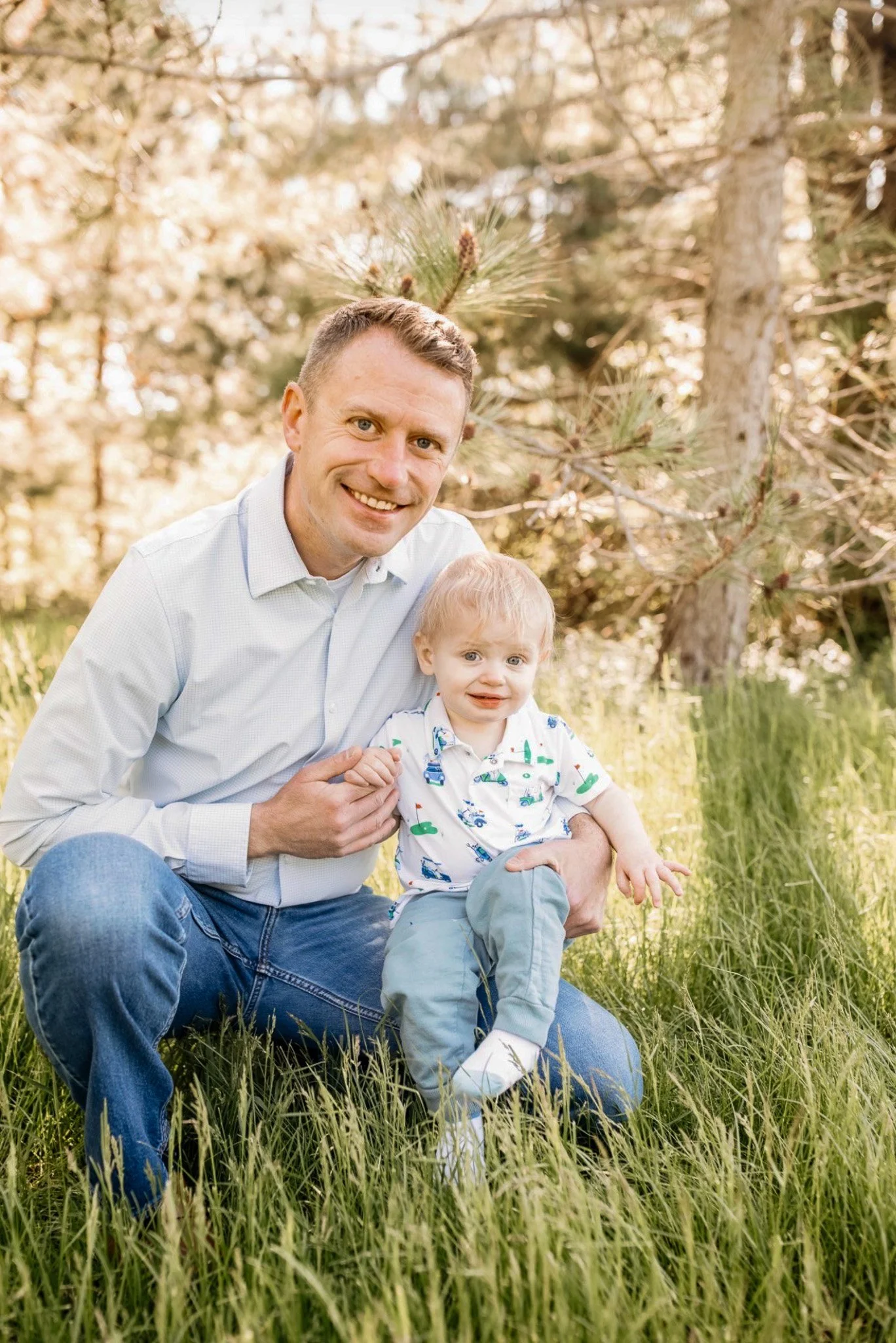 Brent Jacobson and his son sitting on grass in a wooded area, smiling at the camera, surrounded by trees and sunlight.