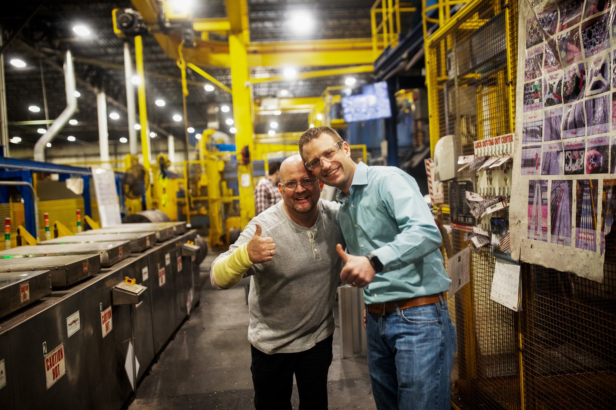 Brent Jacobson smiling and giving thumbs-up inside an industrial warehouse, surrounded by yellow machinery, metal conveyor belts, and shelves with printed notices and items.