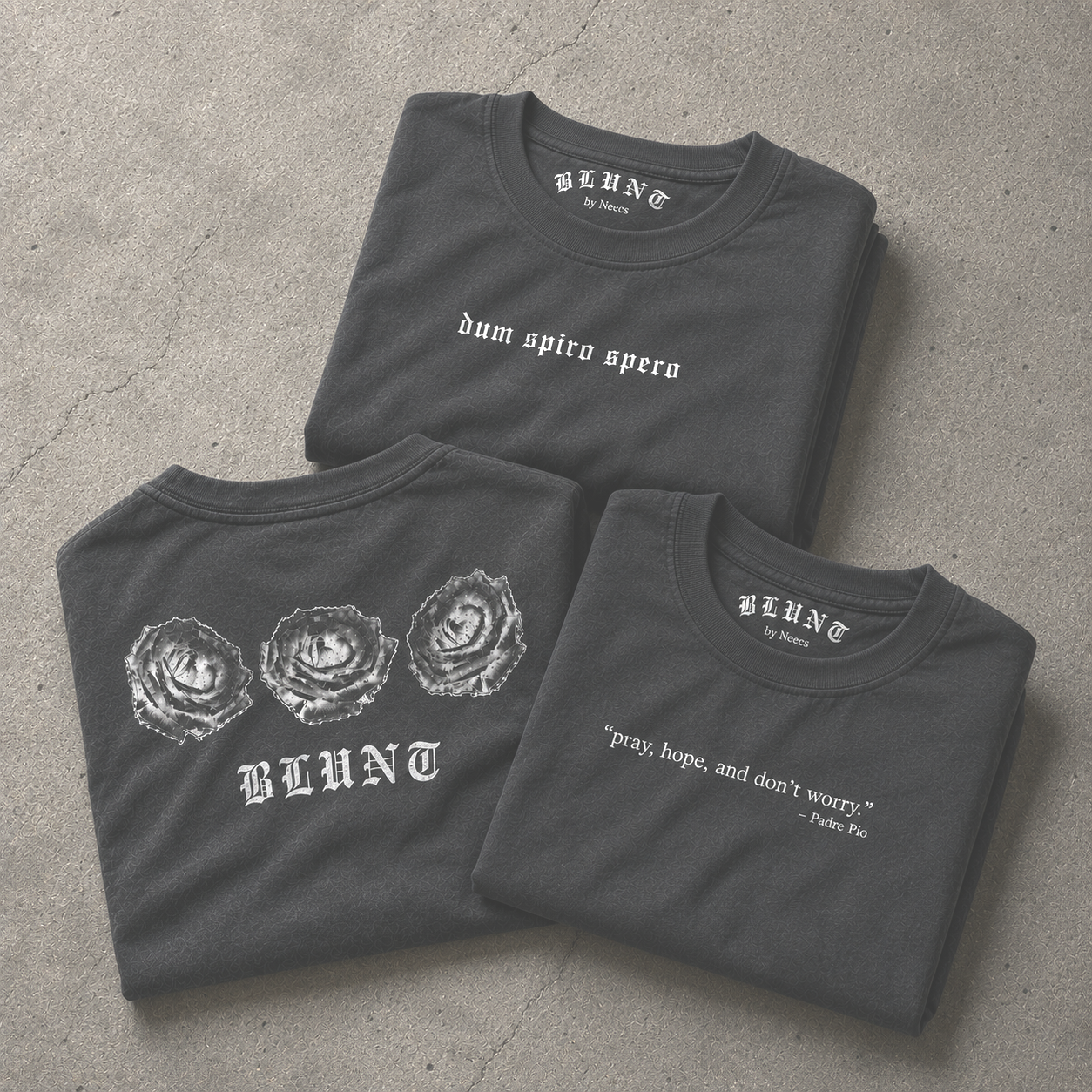 Three folded black T-shirts featuring different white graphic and text designs on a concrete surface.