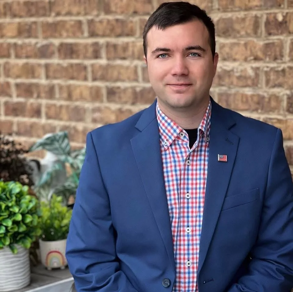 A young man with short dark hair dressed in a blue blazer and a red, white, and blue checkered shirt, standing in front of a brick wall with potted plants on a table beside him.