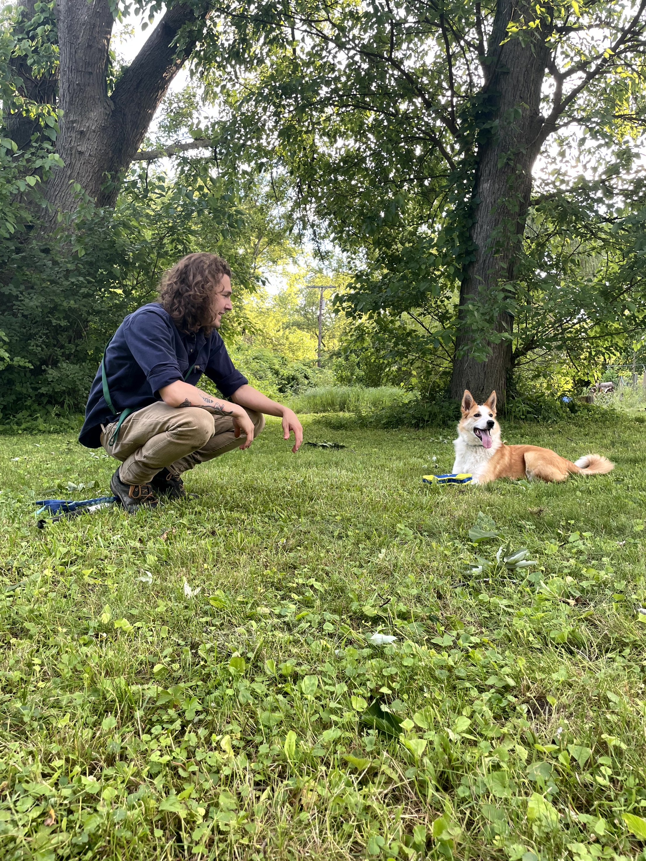 A man with curly hair squatting in a grassy area outdoors, smiling at a sitting dog with its tongue out, in front of tall trees and lush greenery.
