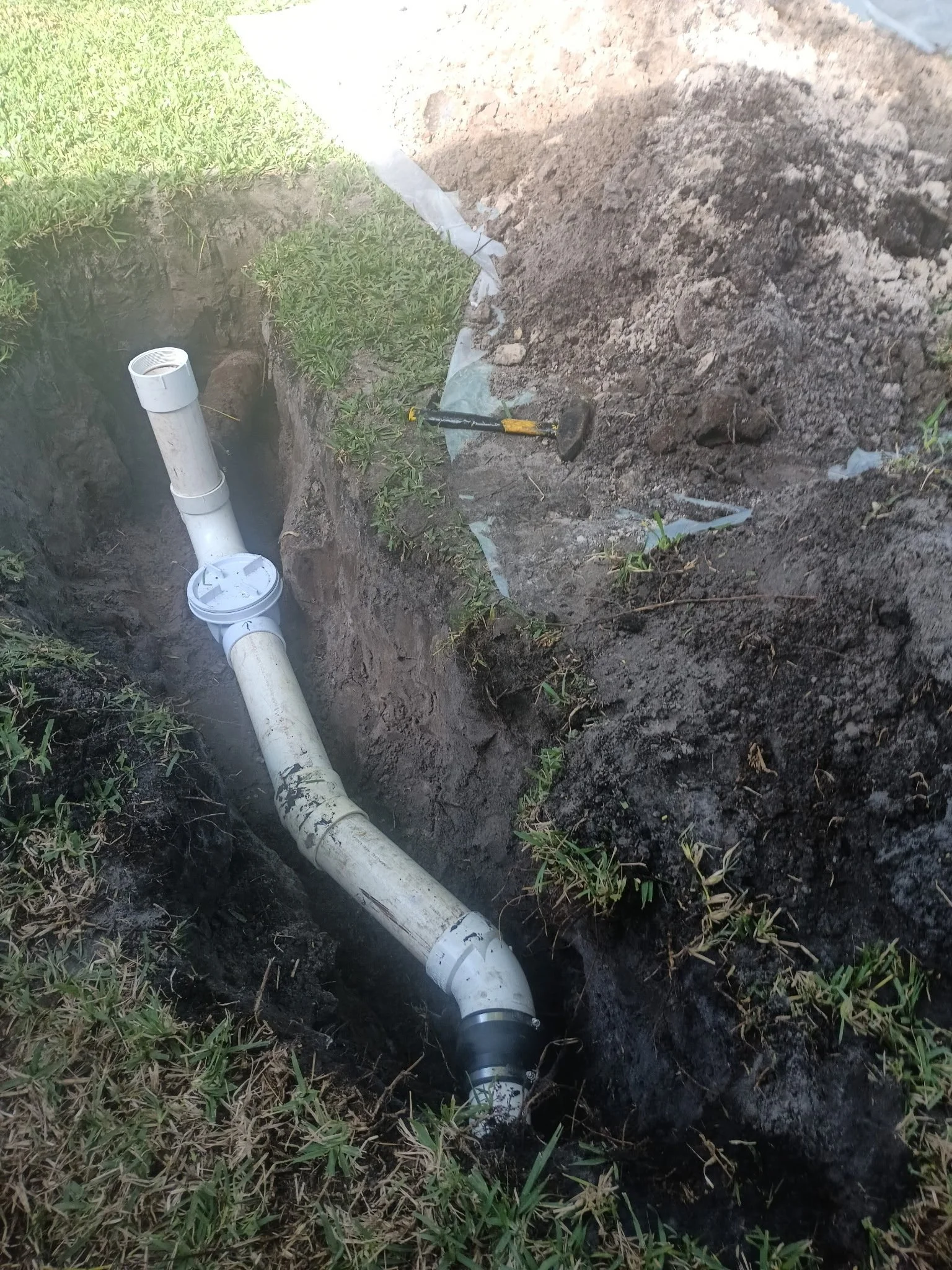 A pipe buried in the ground with soil and grass around it, with a section of the pipe exposed and a pipe wrench lying nearby.