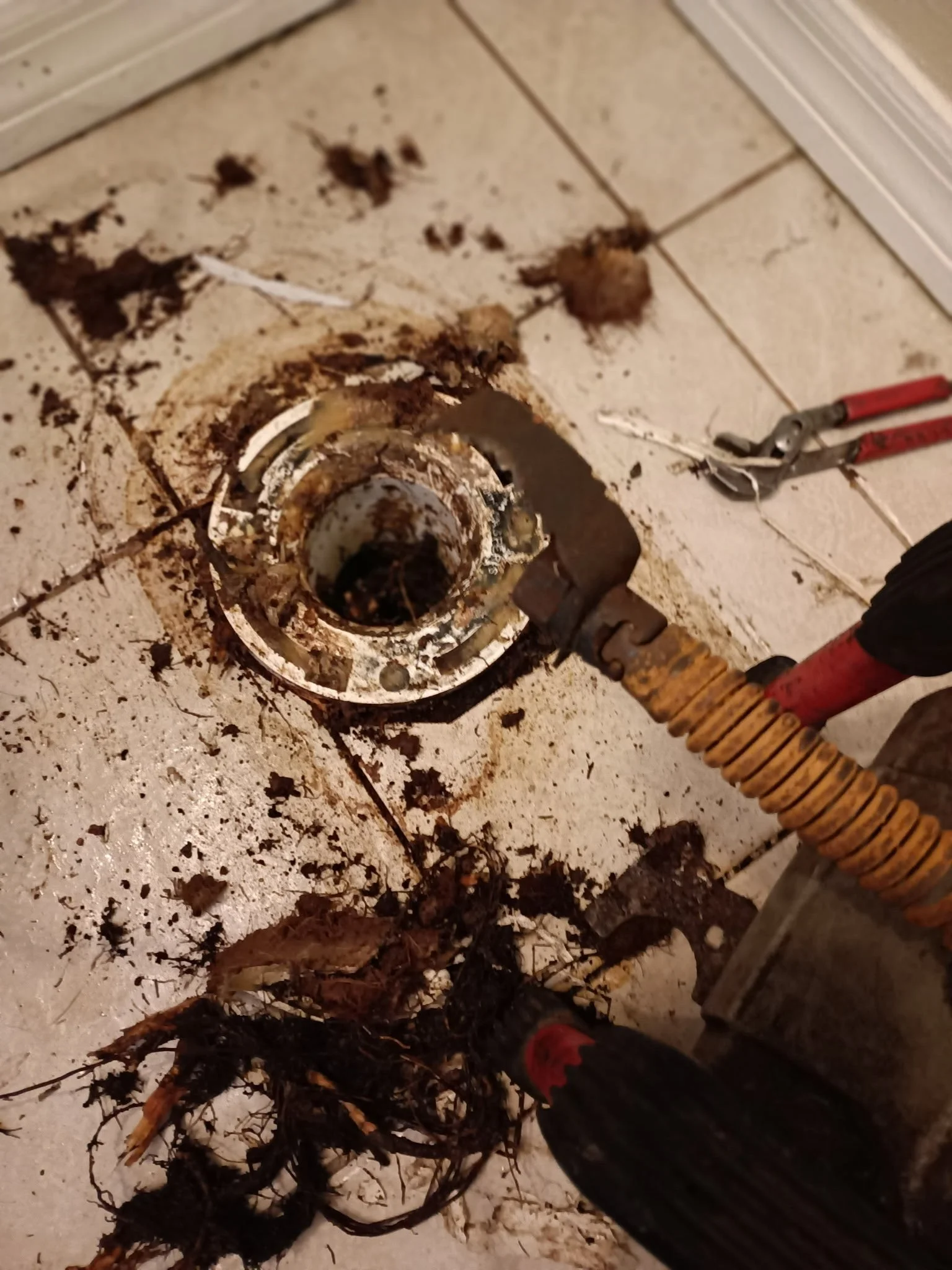 Person using a tool to remove a clogged, rusted metal drain pipe from a tiled floor, with dirt and debris around.