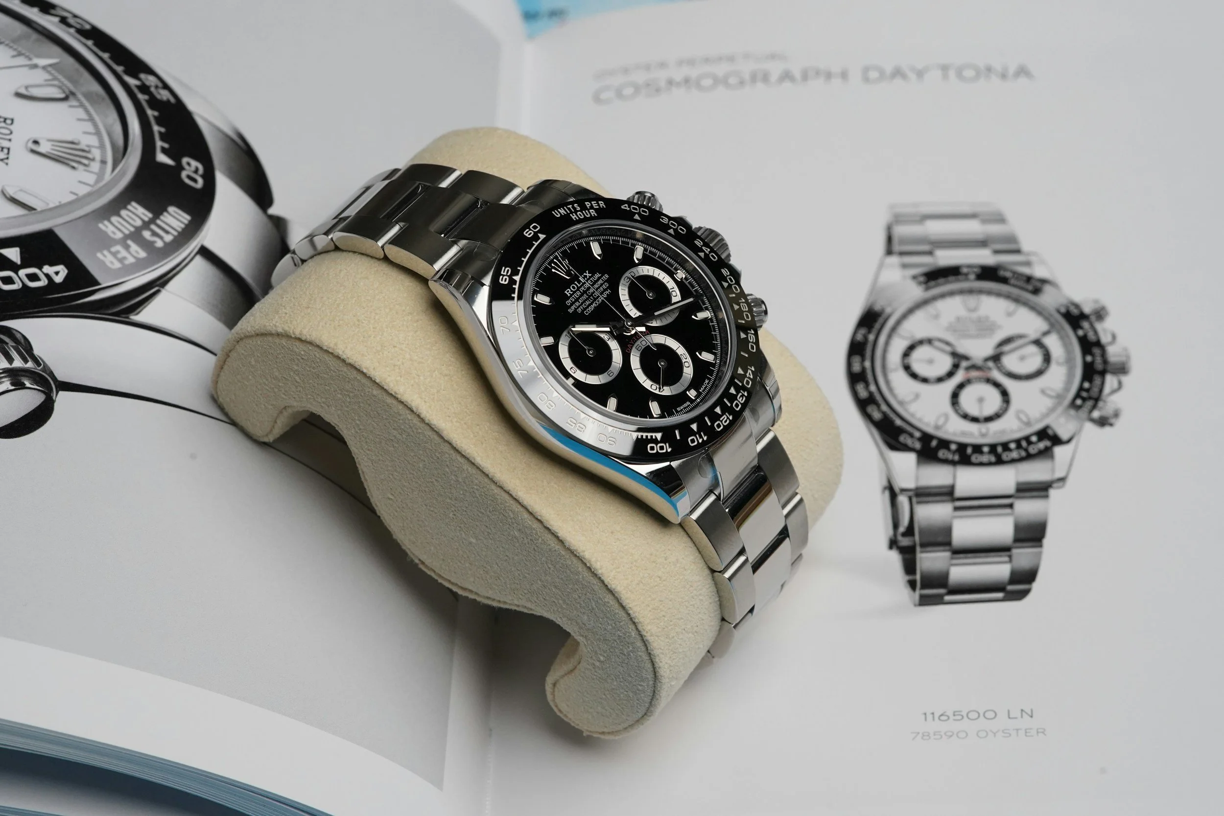 A luxury Rolex watch with a black dial, silver bracelet, and chronograph features, displayed on a beige watch holder with an open booklet showing images of other watches.