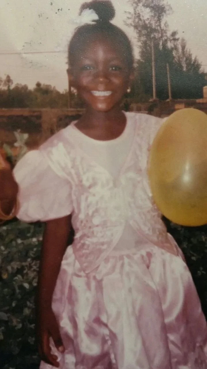 A young girl smiling outdoors, holding a yellow balloon, wearing a light pink dress with puffed sleeves and embroidered details, with trees and electrical poles in the background.