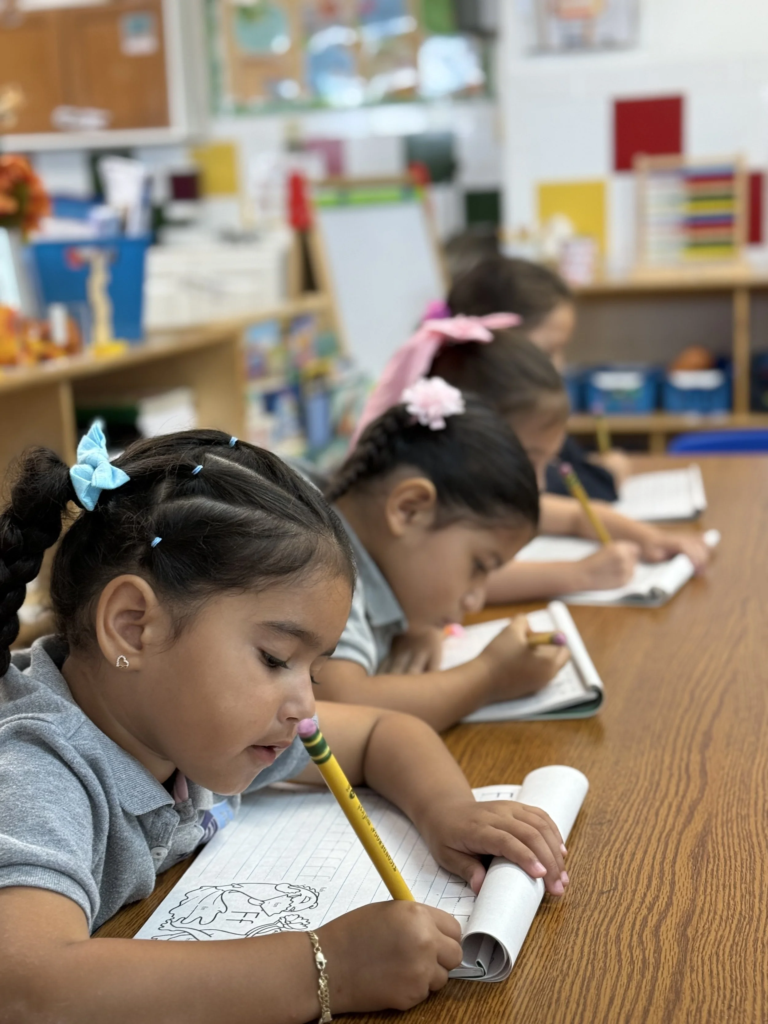 A group of young children, primarily girls, sitting at a wooden table in a classroom, focused on drawing and coloring on paper, with some using pencils and others with coloring tools.