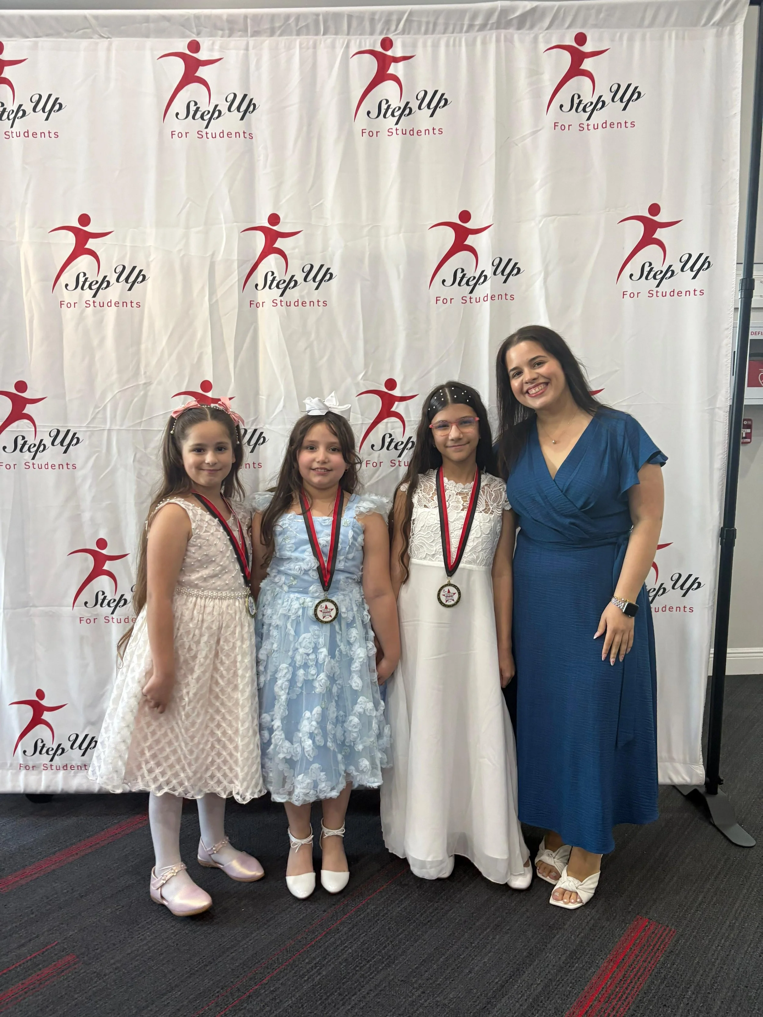 Four females, three young girls and one adult woman, posing at an event with a white backdrop featuring the 'Step Up For Students' logo. The girls are wearing dresses and medals, the woman is wearing a blue dress and beige heels.