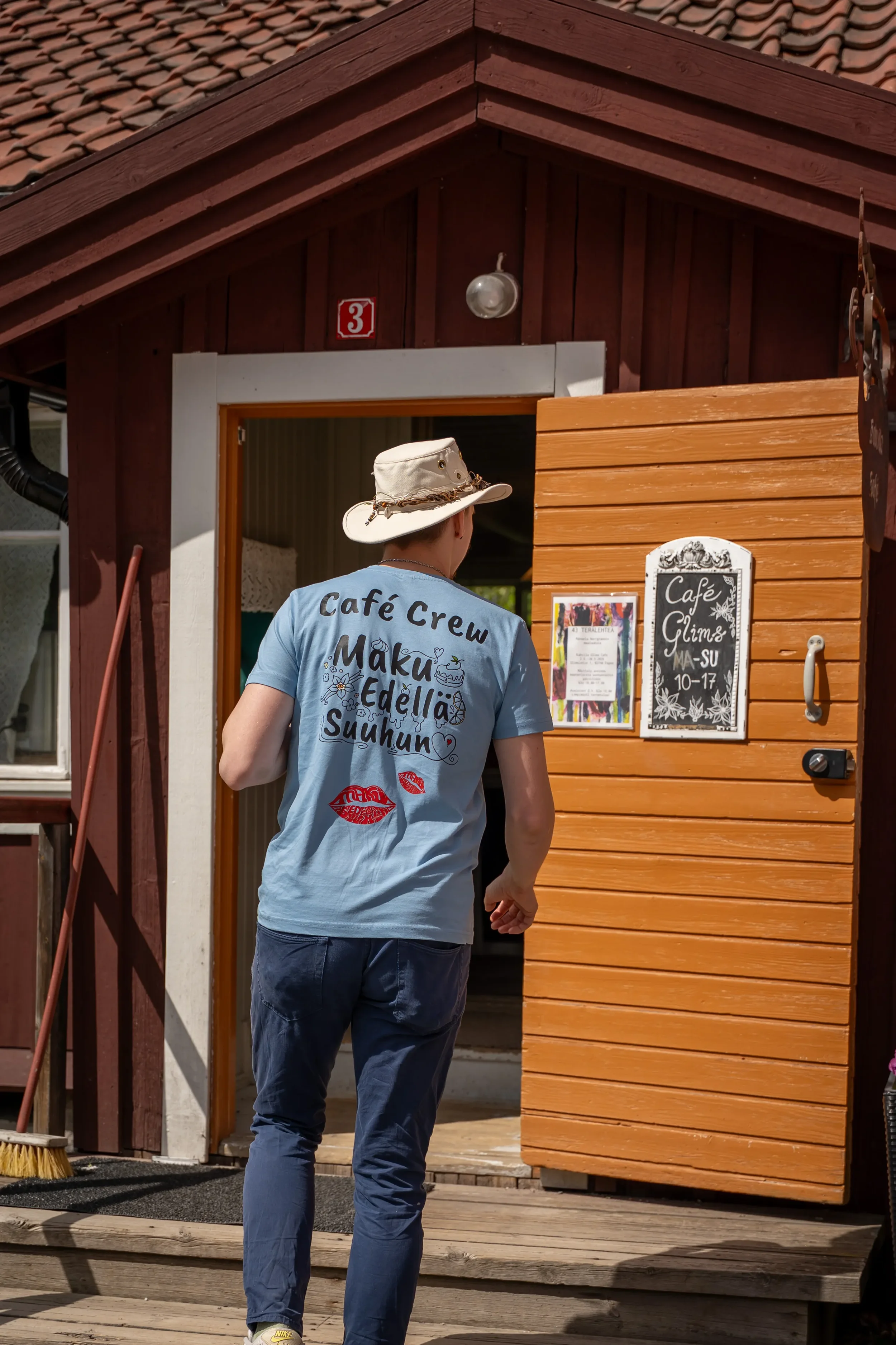 Kahvila Glims Café työntekijä menossa kahvilaan etuovesta. Hänellä on päässään valkoinen hattu, ja hän pukeutuu vaaleansiniseen Cafe Crew T-paitaan ja Tumman sinisiin housuihin.