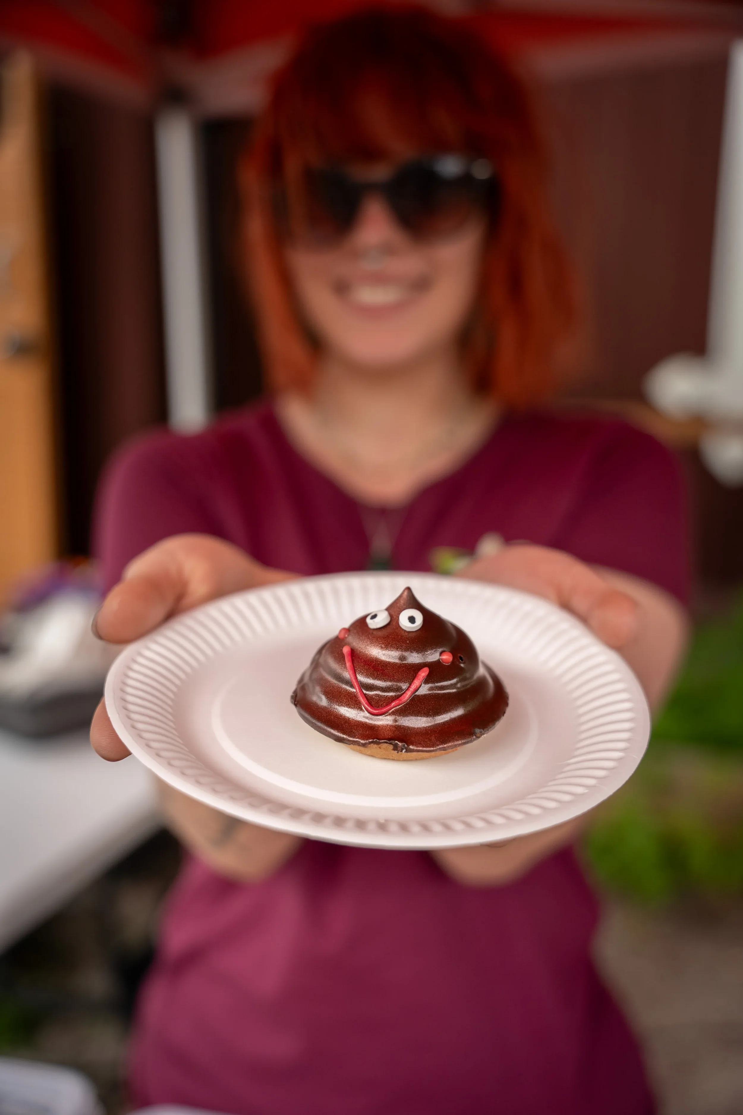 Kahvila Glims Café työntekijä hymyilee ja tarjoaa suklaamuffinia, joka on koristeltu kasvomaisella ja silmillä, kiiltävän suklaakuoren alla, kädessä valkoisella lautasella.