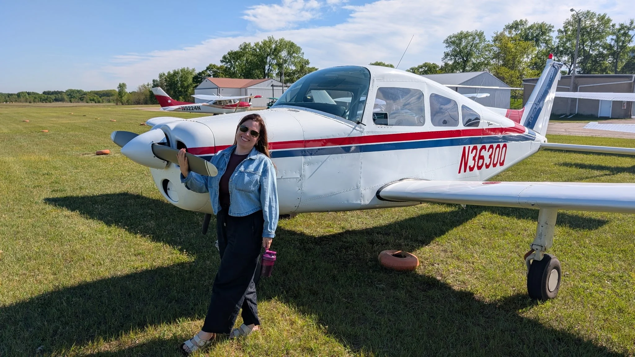 A woman smiling and wearing sunglasses, standing in front of a small white and red airplane on a grassy airfield. She is holding a tablet and wearing a denim jacket, black pants, and sandals. There is another small airplane in the background along with some hangars and trees.