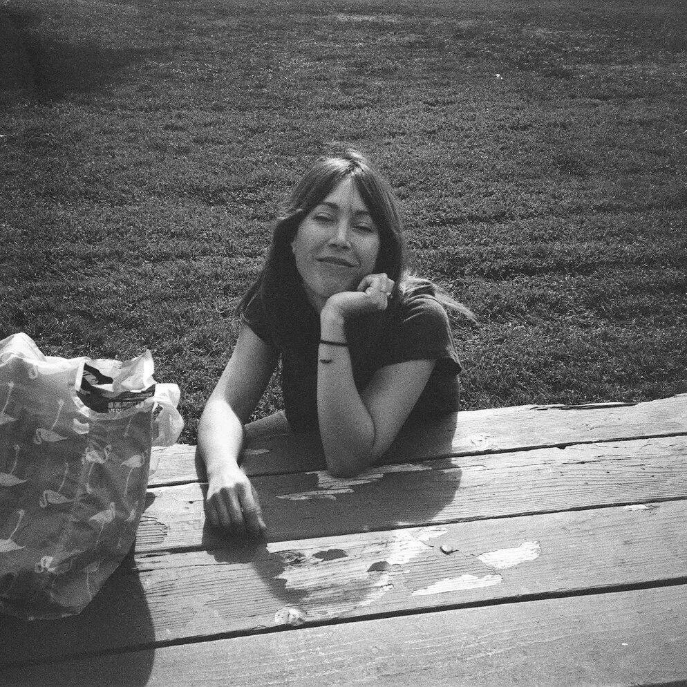 A young woman with long hair resting her chin on her hand at a picnic table outside, with a bag on the table, in black and white photography.