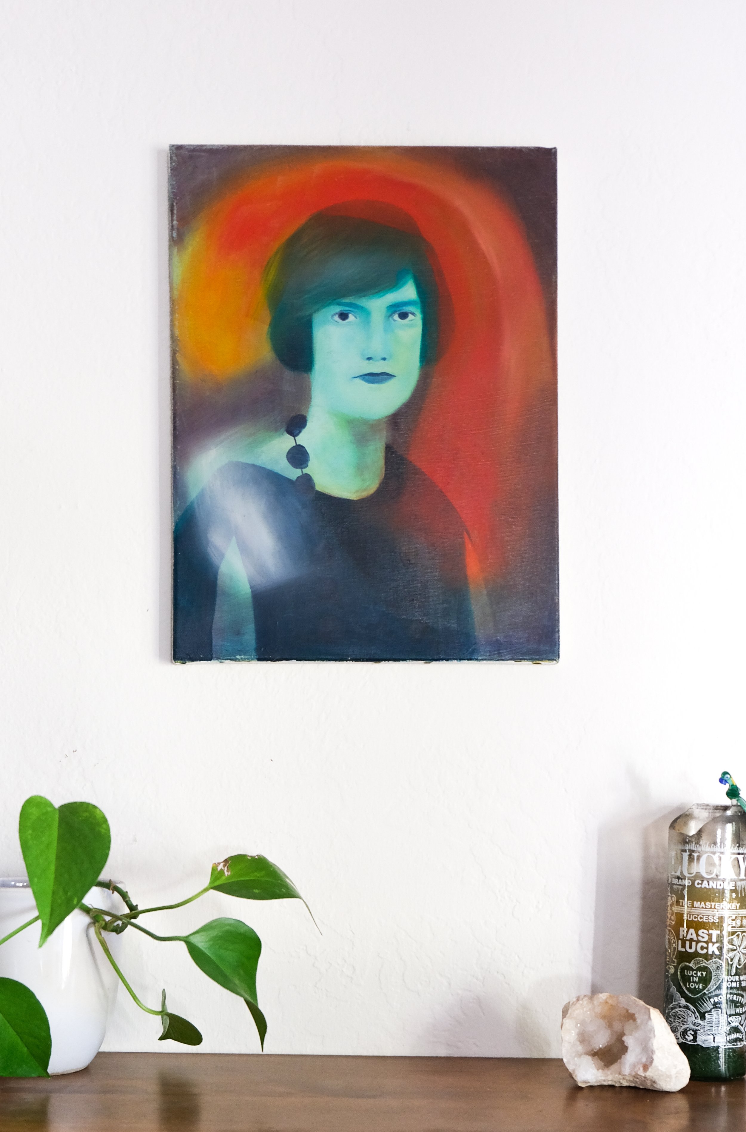 A wall with a colorful abstract portrait painting of a woman with dark hair, a plant with heart-shaped leaves in a white pot, a large quartz crystal, and a container with a Lucky in Love themed candle.
