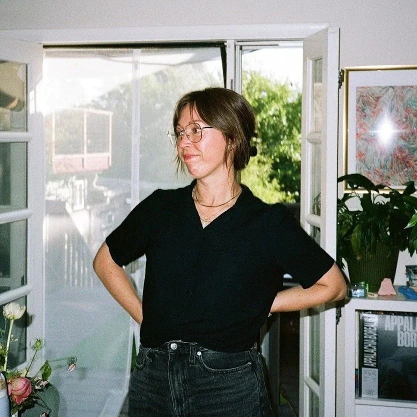 A woman with short brown hair and glasses, wearing a black shirt and dark jeans, standing indoors near open windows, with a background of greenery outside.