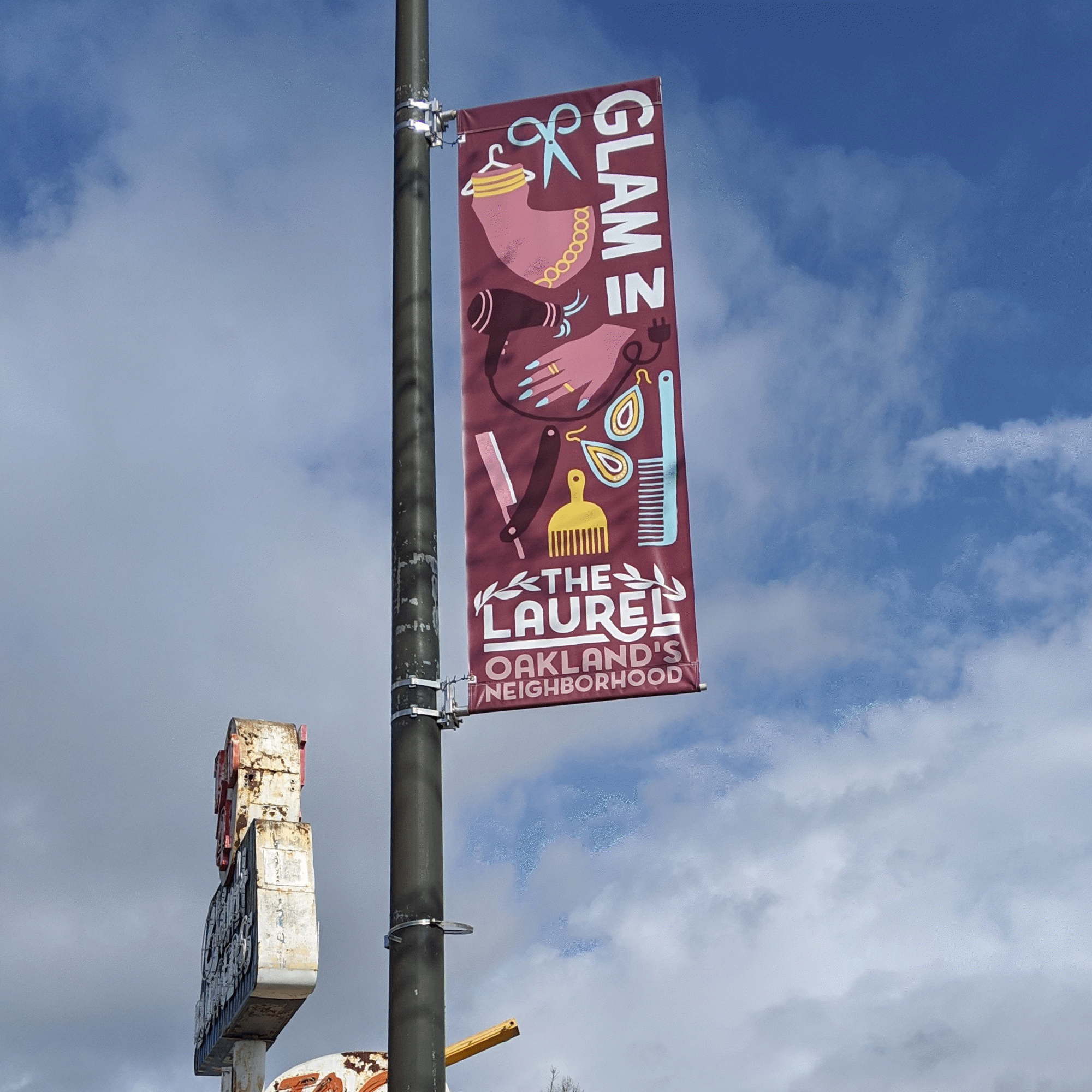 A vertical banner attached to a pole, featuring illustrations of a hairdresser's scissors, a hairdryer, and other hairdressing tools, with text referencing a local neighborhood event or promotion, against a cloudy sky background.