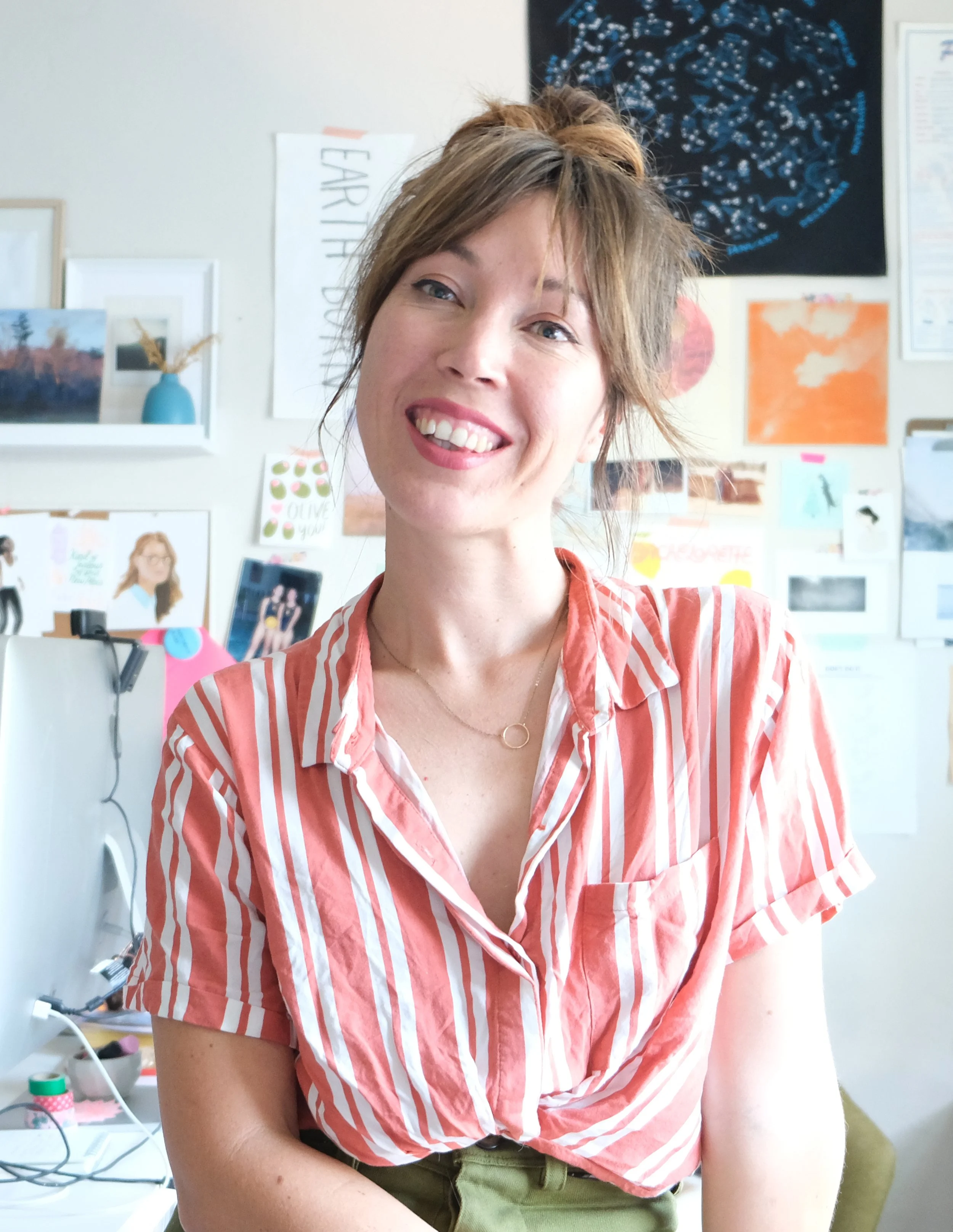 A woman with brown hair and green eyes smiling at the camera, wearing a red and white striped shirt, sitting in an office or creative space with various pictures, posters, and artwork on the wall behind her.