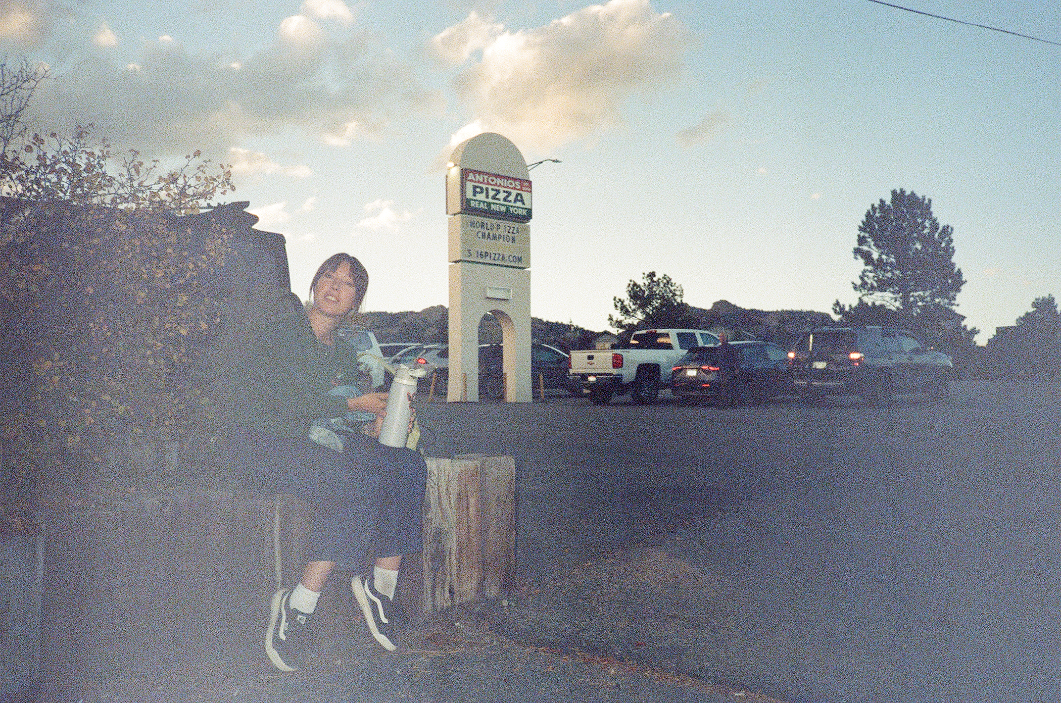 A person sitting on a wooden post at an outdoor location, holding a water bottle, with a parking lot and a sign for Anthony's Pizza in the background, under a partly cloudy sky.