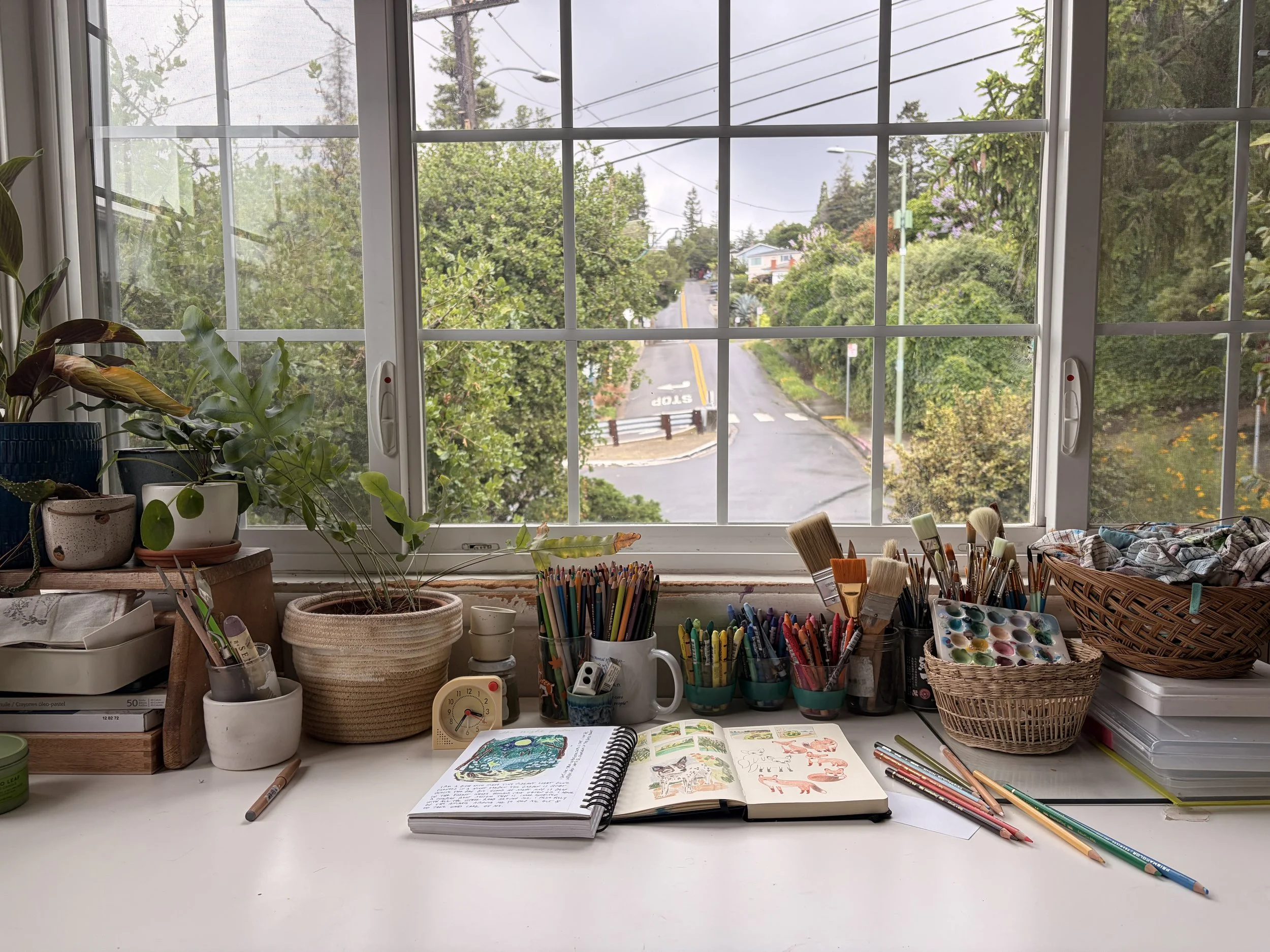 An art workspace with a wide windowsill filled with potted plants, jars, and art supplies like colored pencils, brushes, and a watercolor paint set. An open sketchbook with colorful drawings lies on the white desk, which also holds a small timer and various art tools. Outside the window, a suburban street with trees, houses, streetlights, and power lines is visible under a cloudy sky.
