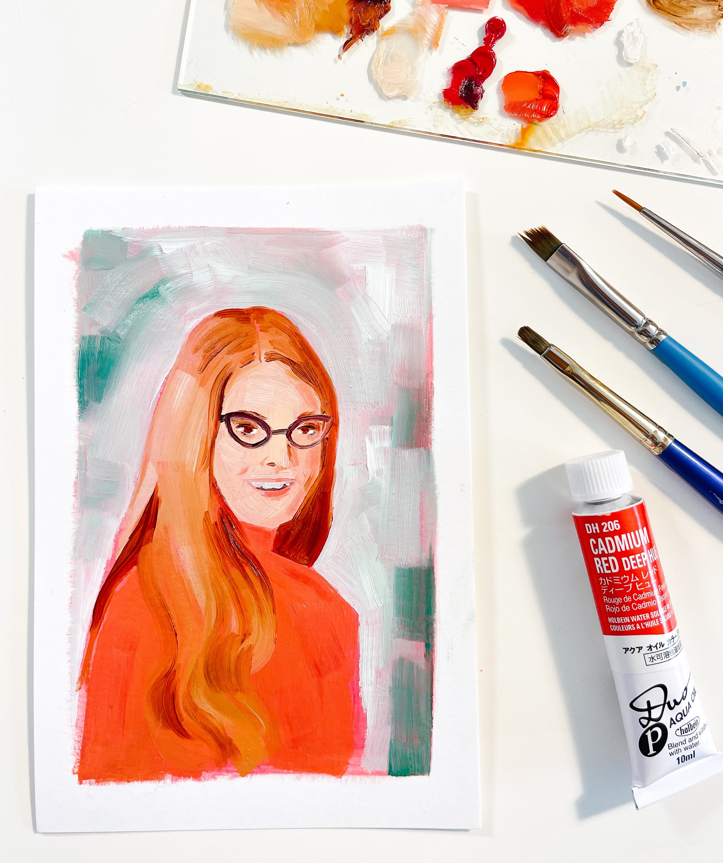A colorful painting of a woman with red hair and glasses, in progress on a white piece of paper, with various brushes and red cadmium hue pigment tube nearby.